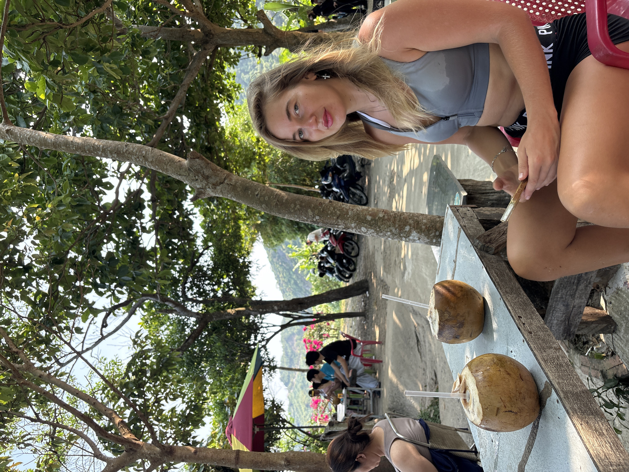 A medium shot depicts a young woman sitting at a table under trees, likely in Đà Nẵng, Vietnam, based on the tropical vegetation and general atmosphere. The woman has light skin, blonde hair, and is wearing a grey sports bra and black shorts with "PINK" printed on them. She is holding what appears to be a vaping device or a similar object. Two opened coconuts with straws are on the table in front of her. In the background, there are other people seated at tables, along with parked motorbikes and green hills. The scene is shaded by trees, suggesting an outdoor cafe or refreshment area.