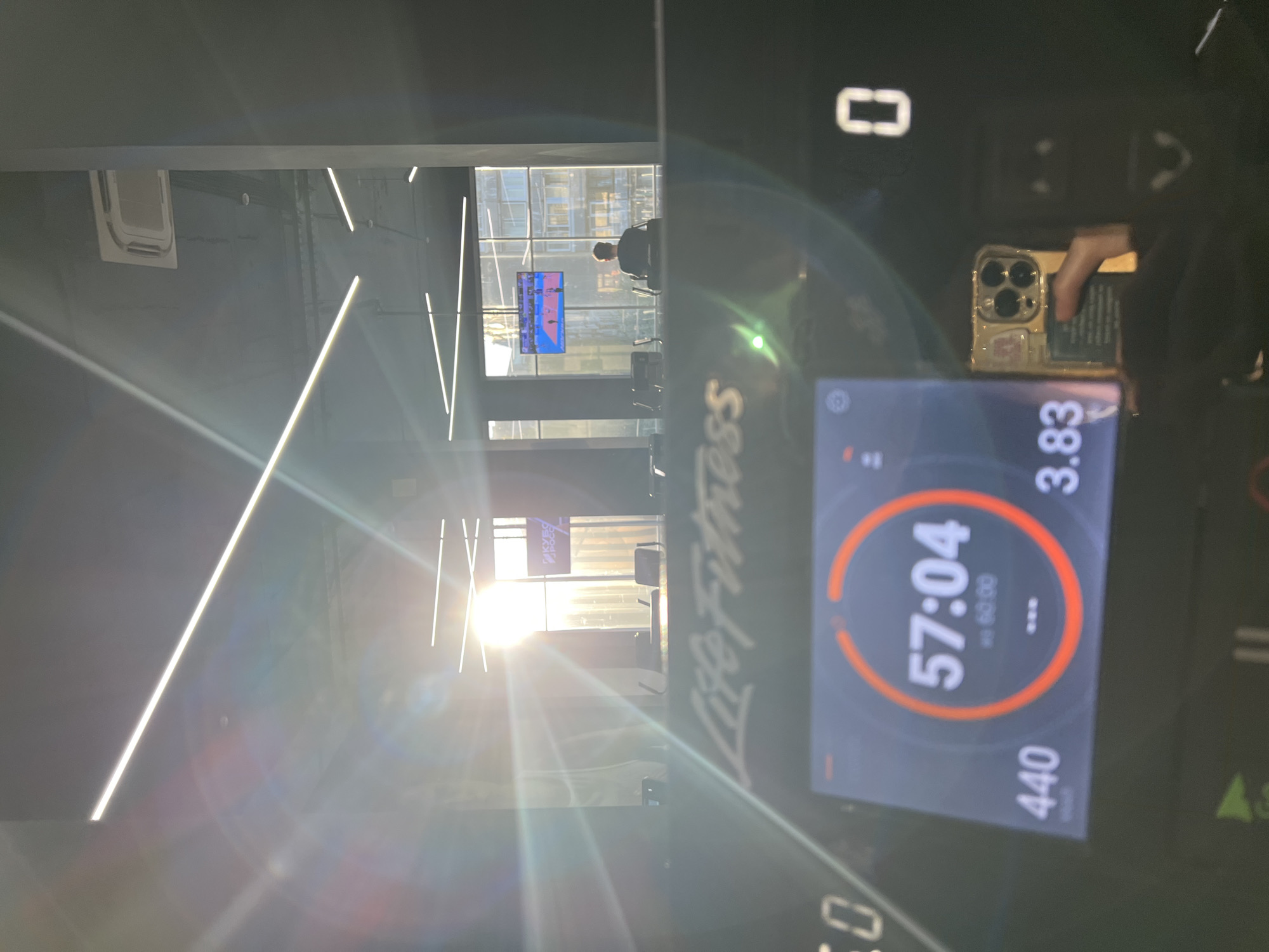 The image shows a view through a window from what appears to be a treadmill at a Life Fitness facility. A screen shows the time 57:04, distance 3.83 and calories 440. Outside the window are buildings and other treadmills. One person is using another treadmill. Two televisions are broadcasting. The location is Khimki, Russia, as indicated by signage in the window.