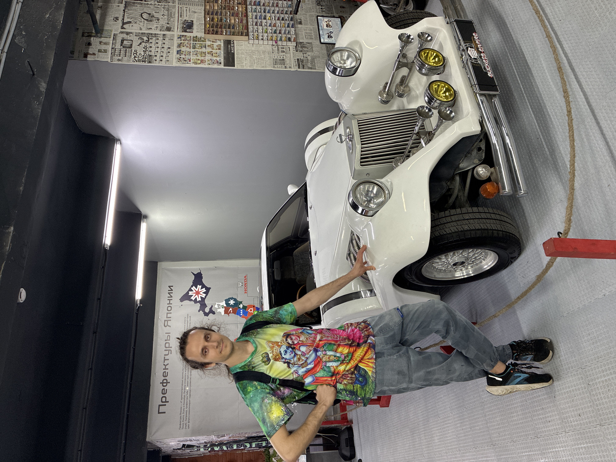 The image shows a man posing next to a white vintage-style car in what appears to be a garage or showroom. The man is wearing a brightly colored shirt with a Hindu deity print, grey jeans, and black sneakers. He has long hair and a backpack slung over his shoulder. He is touching the front of the car. The car has a prominent grill and multiple lights. The background includes posters and newspapers on the walls, a map of Japan with the title "Prefectures of Japan", and a Honda logo. The room is well-lit, suggesting an indoor setting. The location is likely Moscow, Russia, based on the Cyrillic text on the posters and map.