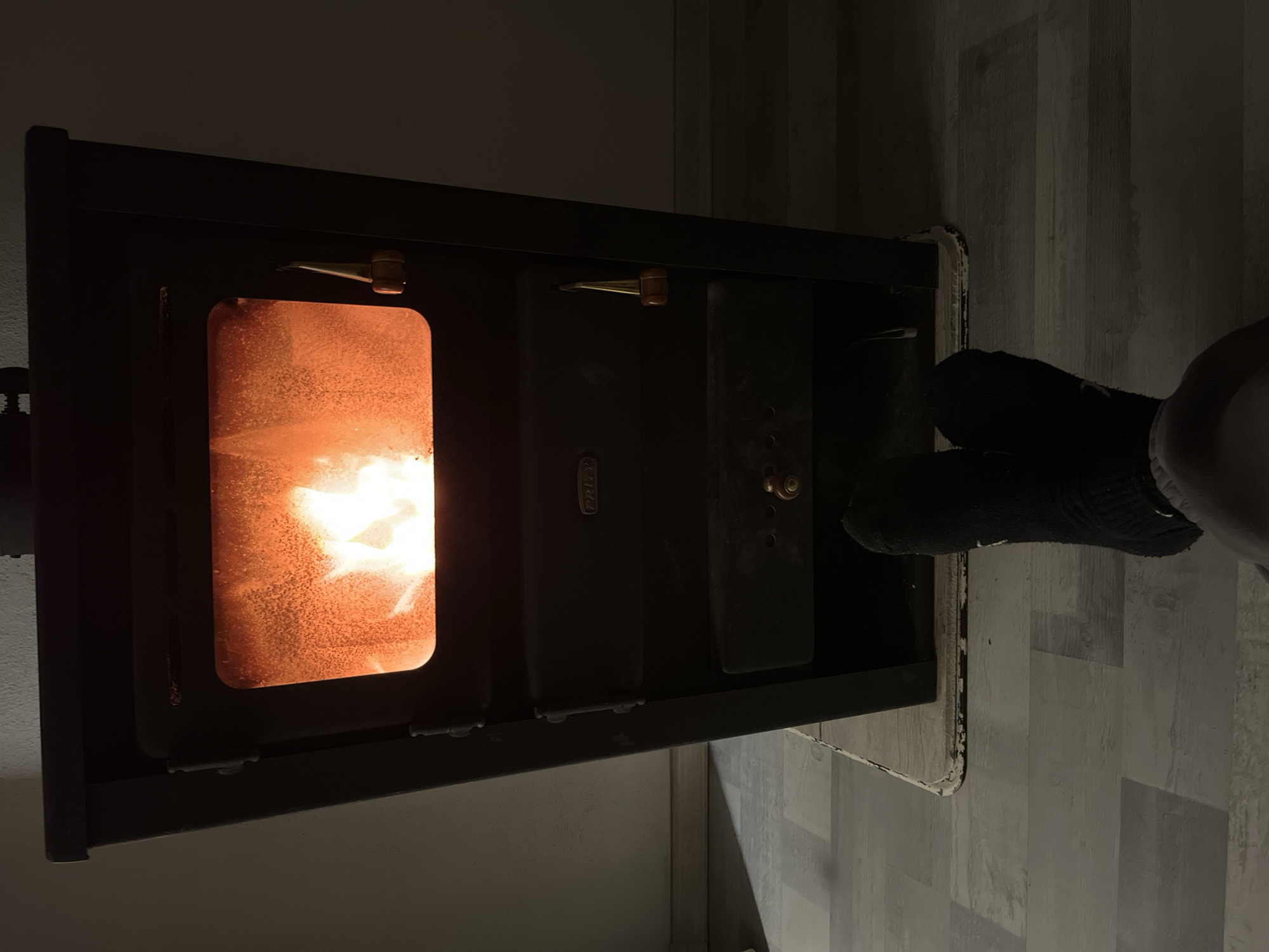The image shows a black wood-burning stove with a fire visible through the glass door. The fire is bright orange and white. The stove is located in a room with a floor that appears to be covered in grey and white patterned linoleum or laminate. The room is dimly lit, with the fire providing the primary light source. A person's leg and foot, clad in a black sock, are visible to the right of the stove. The location is not identifiable as Karavelovo, Bulgaria, based on the provided image.