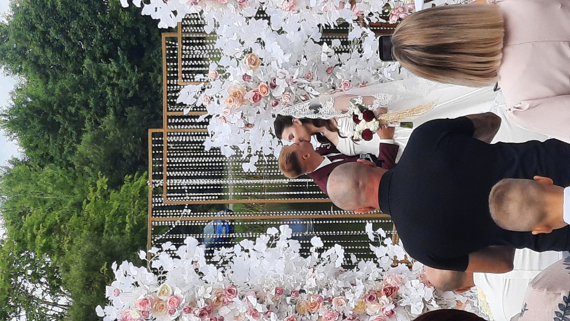 A wedding ceremony is taking place outdoors, likely in the vicinity of Жодзішкаўскі сельскі Савет, Belarus, given the provided context. A bride and groom are embracing in a kiss under a decorative archway adorned with white and pink flowers and shimmering crystal strands. The groom, wearing a burgundy suit, is kissing the bride, who is in a white wedding dress and veil, holding a bouquet of red and white flowers. In the foreground, the backs of several guests are visible, including a man in a black polo shirt, a child with short blond hair, and a woman with short blonde hair wearing a pale pink dress. Behind the couple, a body of water and trees can be seen, with a blue car parked near the water's edge. The environment suggests a park or garden setting.
