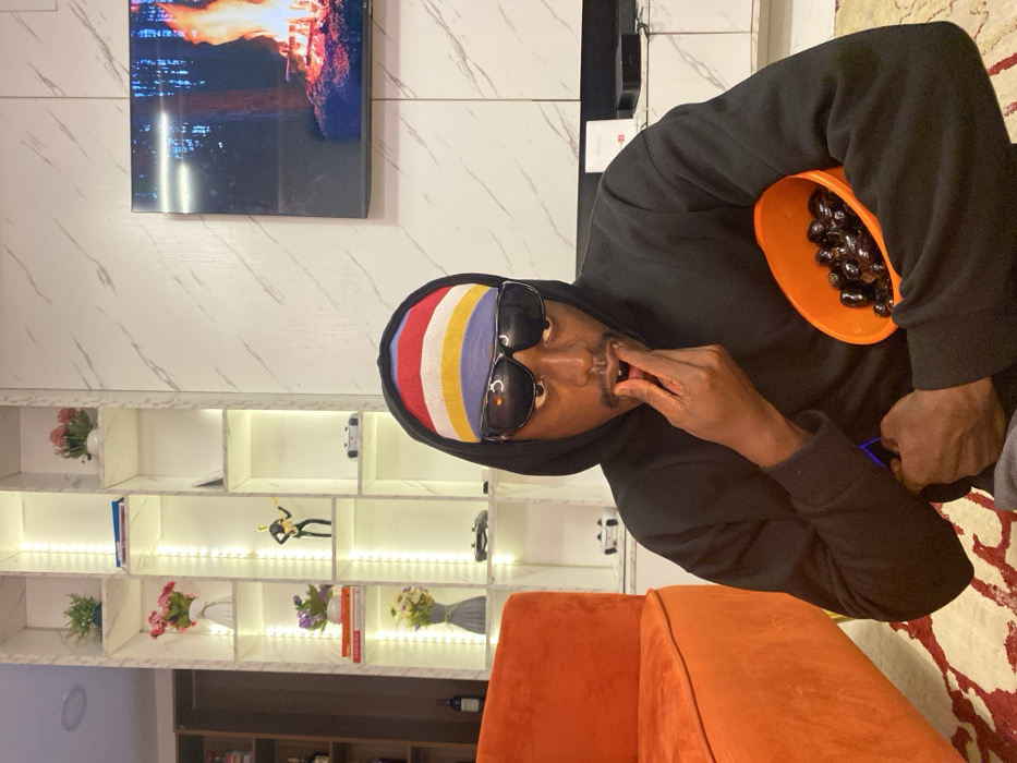 A man wearing a black hooded sweatshirt, sunglasses, and a colorful striped head covering is eating olives out of an orange bowl. He has a mustache and is dark-skinned. In the background, there is a modern-looking wall with a TV, a lit-up display shelf with vases, flowers, and decorative items. There is an orange sofa or chair in the foreground, and a patterned carpet is visible on the right side of the image.