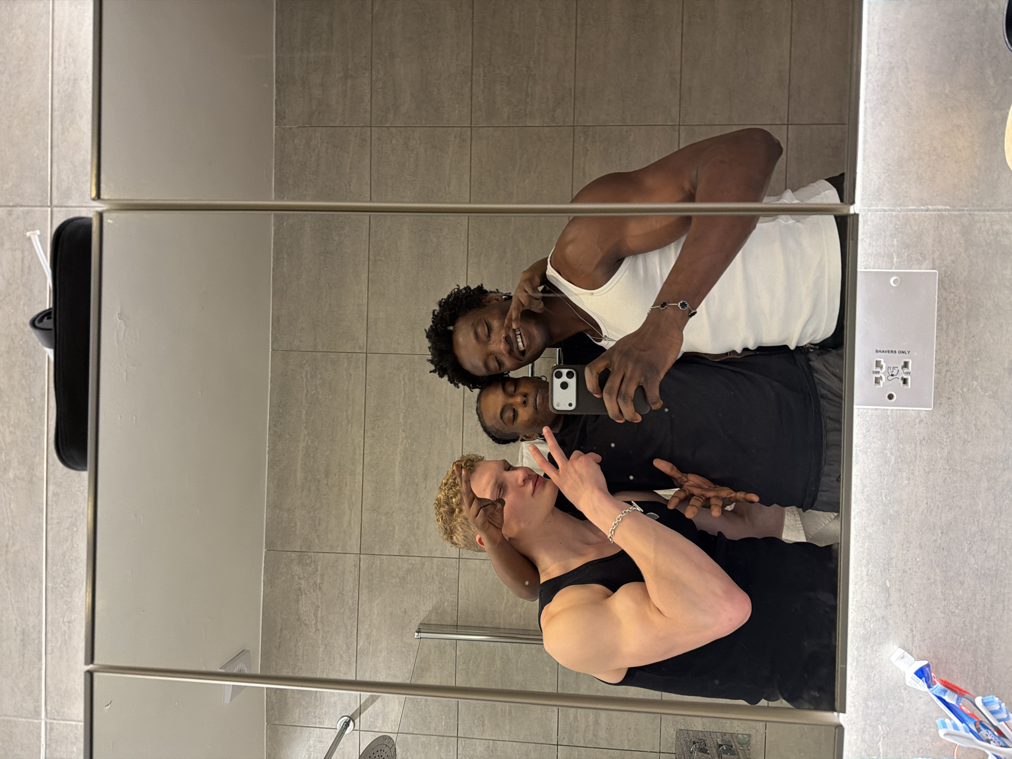 Three individuals pose for a mirror selfie in a bathroom setting. The bathroom features grey tile walls and a large mirror divided into sections. Toiletries, including toothbrushes and toothpaste, are visible in the lower right corner. One individual, positioned closest to the mirror, is light-skinned with curly blonde hair, wearing a black tank top and sporting a silver bracelet. The second individual is dark-skinned, wearing a black shirt, and holding a smartphone to take the photo. The third individual is also dark-skinned, wearing a white tank top, and is partially embracing the second individual. The location is likely a bathroom somewhere in the city of London, United Kingdom.