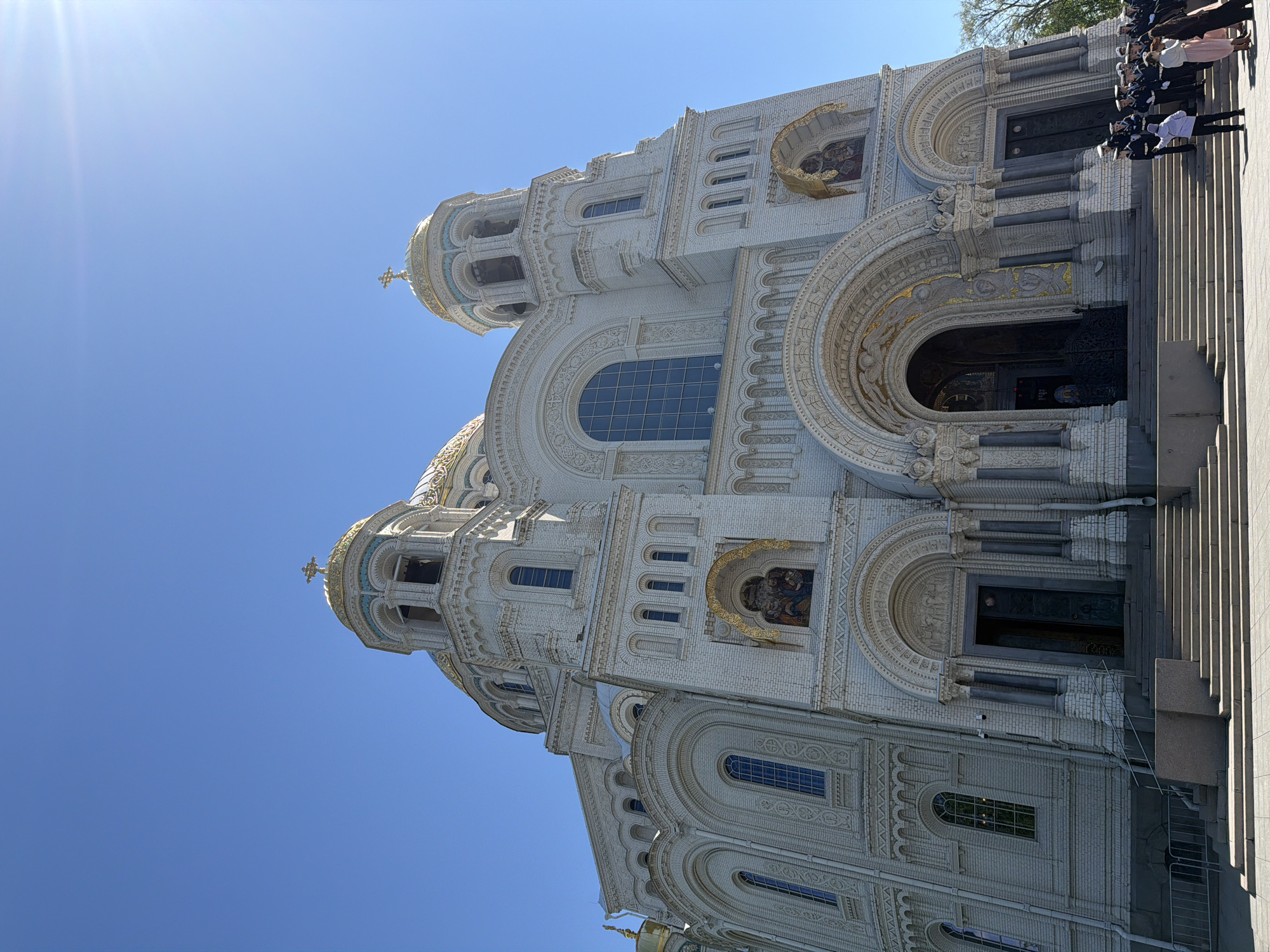 The image shows the Naval Cathedral of Saint Nicholas in Kronstadt, Russia. The cathedral is a white brick building with golden domes and crosses on top. The architecture includes arched windows and doors, intricate carvings, and a mosaic of saints. People in naval uniforms stand near the entrance, possibly participating in a ceremony. The sky is blue, and sunlight shines brightly.
