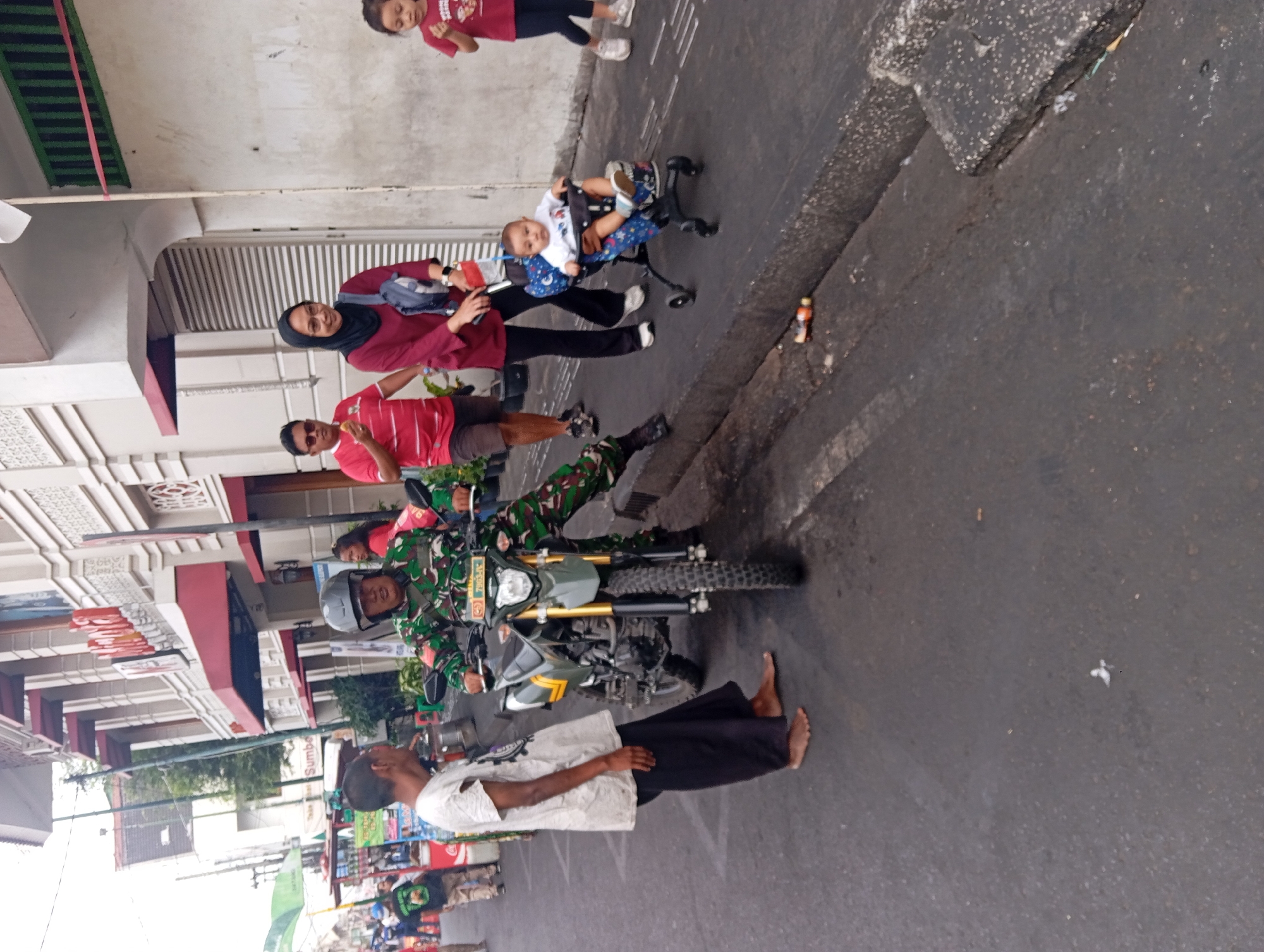 The image captures a street scene in the city of Yogyakarta, Indonesia, likely in a busy area with shops and pedestrian activity. In the foreground, a person in military camouflage attire is seated on a motorcycle, appearing to be interacting with a barefoot individual in a white t-shirt and dark pants standing next to the bike. To the right, a woman in a maroon hijab and a man in a red and white striped shirt walk past, the woman pushing a stroller with a baby inside. Further in the background, other individuals and a food cart are visible, indicating a lively urban environment. The buildings lining the street have a colonial architectural style, common in Yogyakarta.