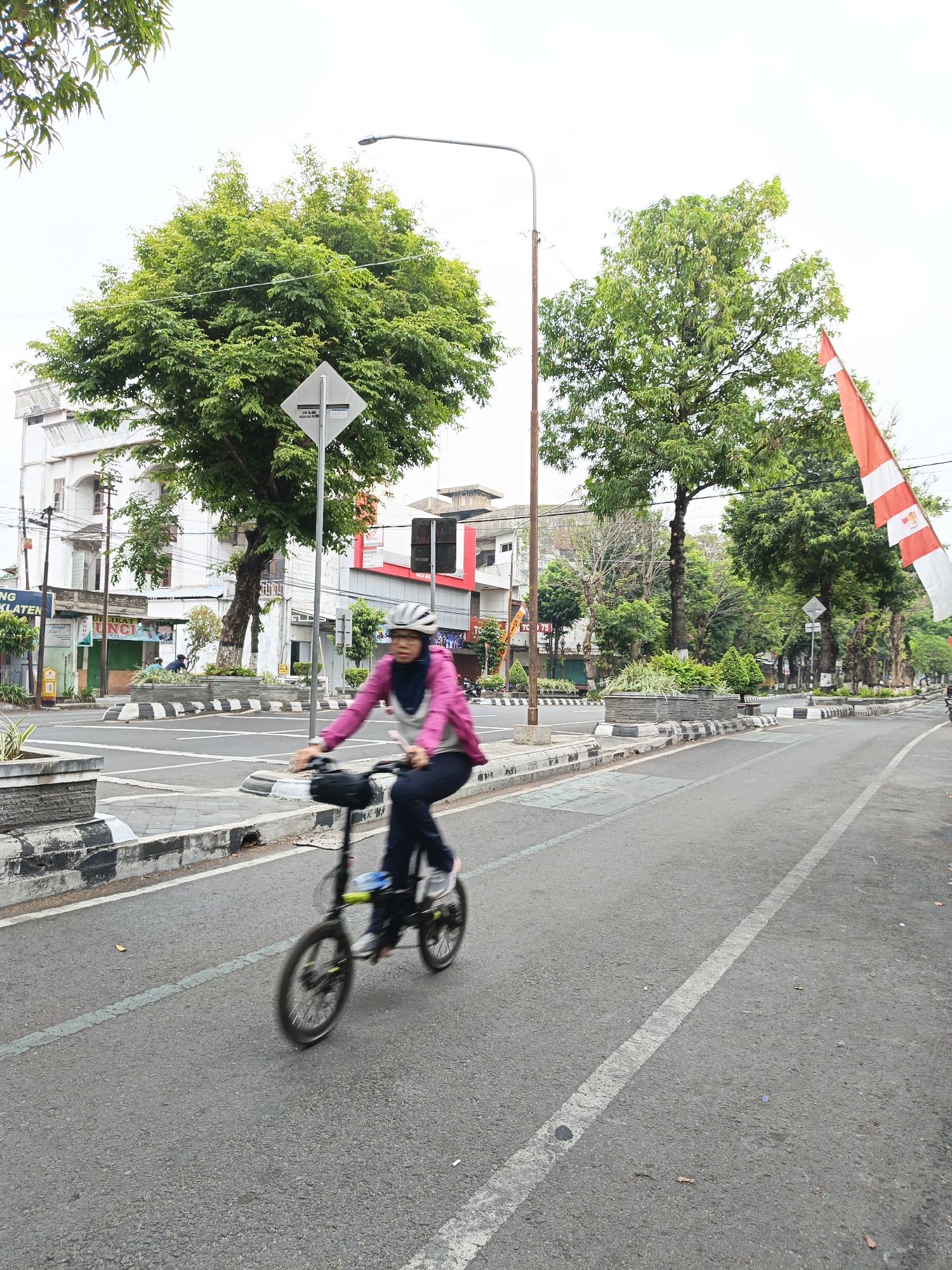 A blurred motion shot captures a woman cycling on a street in Klaten, Indonesia. She is wearing a pink jacket, dark pants, a hijab, and a helmet. The woman is on a folding bicycle, and her body is leaning forward as she pedals. The street is paved with asphalt and has a white line marking the lane. On the left side of the street, there is a sidewalk with a decorative planter and some trees. In the background, there are buildings, streetlights, and more trees. Indonesian flags are visible on the right side of the frame, indicating a possible patriotic event or public display. The sky is overcast.