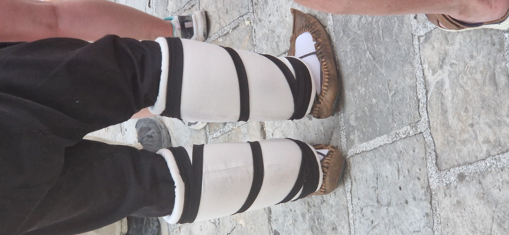 The image shows a close-up view of a person's legs and feet, wearing traditional Montenegrin attire. The person is wearing black trousers, white leg wraps adorned with black straps, and brown leather tsakonia (traditional Montenegrin shoes). The background consists of a cobblestone pavement, characteristic of the old town of Budva, Montenegro. The lighting suggests it is daytime, and the focus is on the lower half of the person's body, highlighting the details of their costume and footwear.