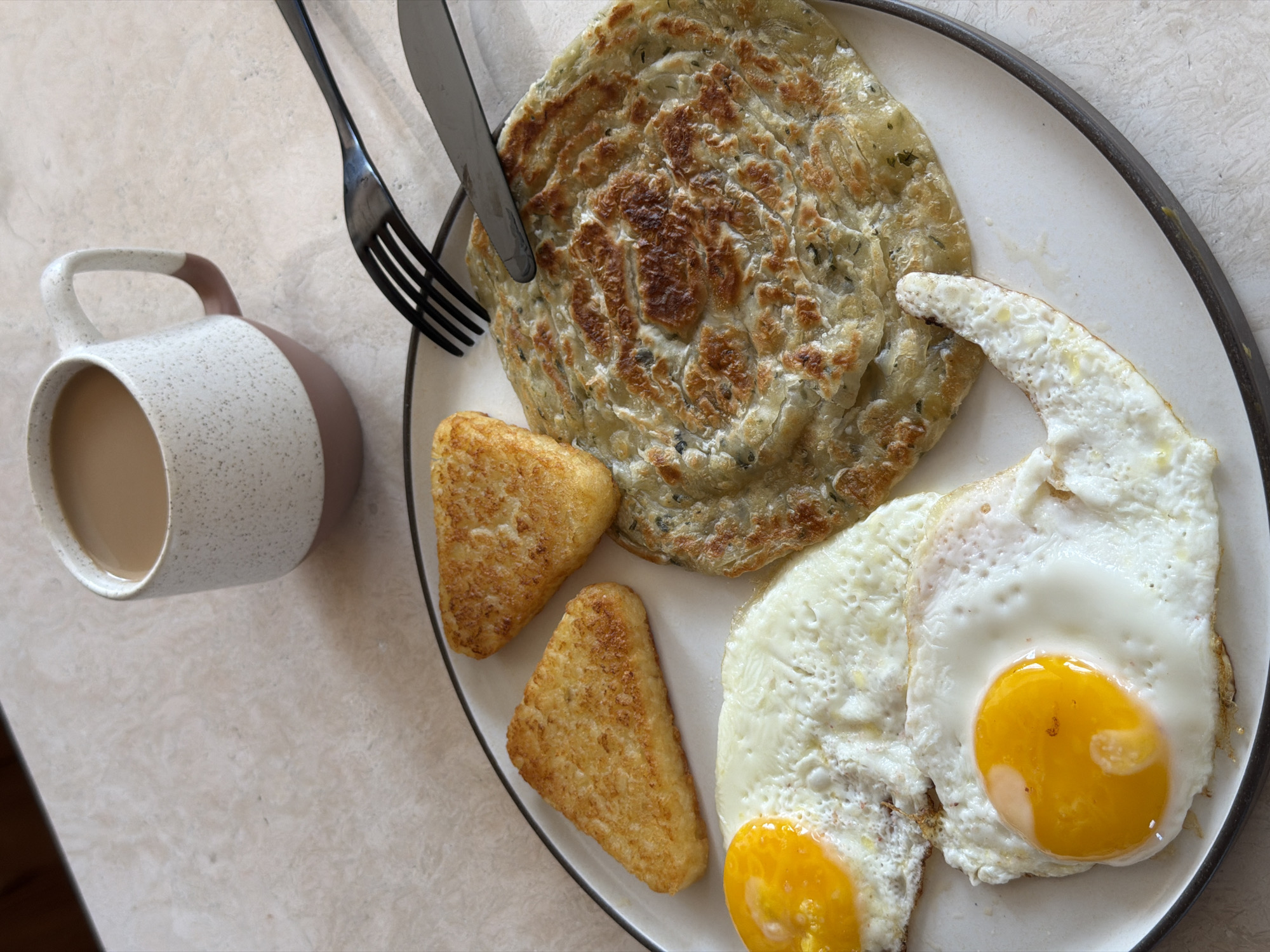 The image depicts a plate of breakfast food alongside a mug of coffee, likely captured indoors on a countertop. The plate contains two fried eggs with bright yellow yolks, two triangular hash browns, and a circular flatbread. A fork and knife rest on top of the flatbread. The mug of coffee is positioned to the left of the plate. The countertop has a light, mottled surface. The location in the city of Mengwi, Indonesia, cannot be verified from the visual content of the image.