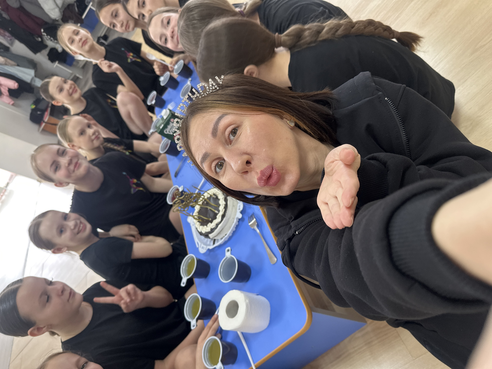 A high-angle selfie shows a woman wearing a "birthday" tiara, with a birthday cake and a group of young girls around a blue table. The scene appears to be indoors, potentially in a dance studio or similar space, with a light-colored wooden floor. Everyone is wearing dark t-shirts and some have their hair neatly styled, possibly related to a dance class. The birthday cake is on the table, along with cups and what seems to be a roll of paper towels. The group is gathered closely around the table for the celebration. Given the presence of the dance-related details like the attire and hairstyles, and considering that the photo includes a cake, the group might be at a birthday party for the woman in the selfie, or for one of the girls in the group. There is nothing in the image to explicitly place the location in Votkinsk, Russia.