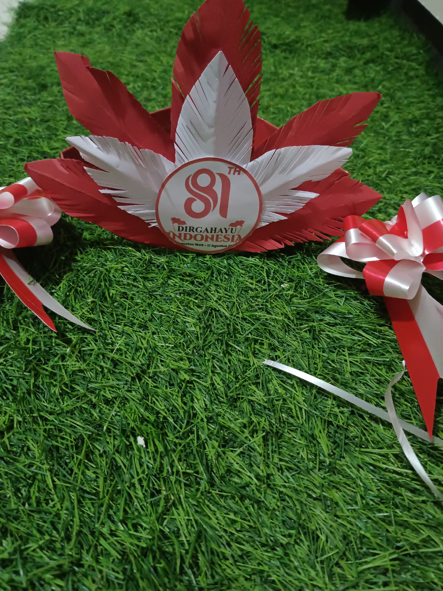 The image depicts a festive, red and white-themed centerpiece resting on artificial green grass. The centerpiece is shaped like a crown or headdress, adorned with paper feathers in red and white. In the center of the headdress is a circular emblem that reads "81 TH DIRGAHAYU INDONESIA 17 AGUSTUS 1945 - 17 AGUSTUS 2026". On either side of the centerpiece are large, decorative bows made of red and white ribbon, with trailing ribbons extending downwards. The lighting suggests an indoor setting, possibly a decorated event space. There is no indication of the city of Kawua, Indonesia, or any specific urban location. The scene is static, with no people or actions depicted.