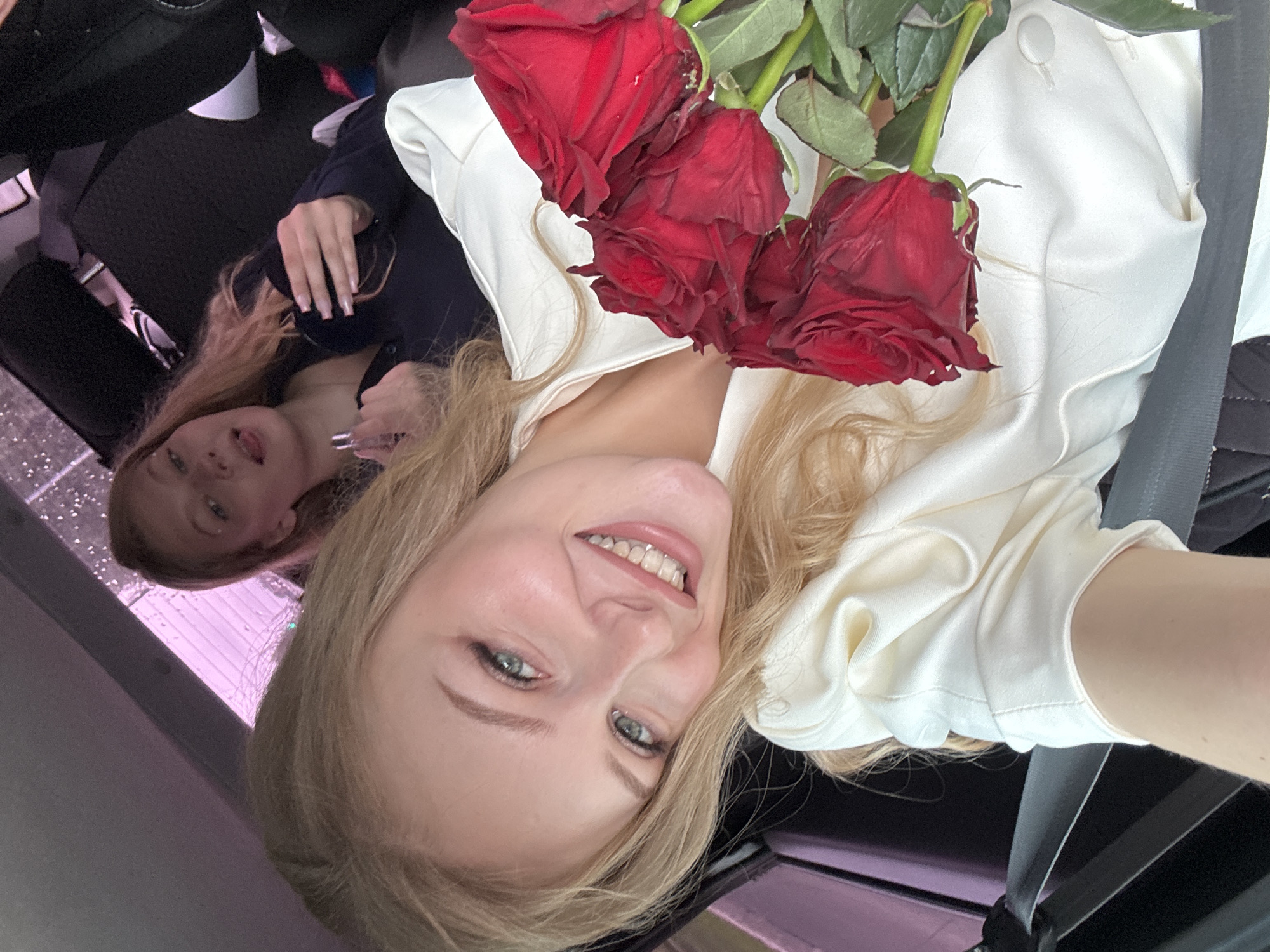 Two young blonde women are in the back seat of a vehicle. The woman in the foreground is closer to the camera and is smiling broadly, holding a bouquet of red roses. She is wearing a white satin shirt. The woman in the background is to her left, with her tongue sticking out, holding a small tube that appears to be lipstick. The interior of the car is visible, including the seats and a seatbelt. The background outside the car windows is blurred and shows a pinkish light, possibly from city lights or a screen. The scene is consistent with being in the city of Perm, Russia, where such vehicles and lighting conditions are common.