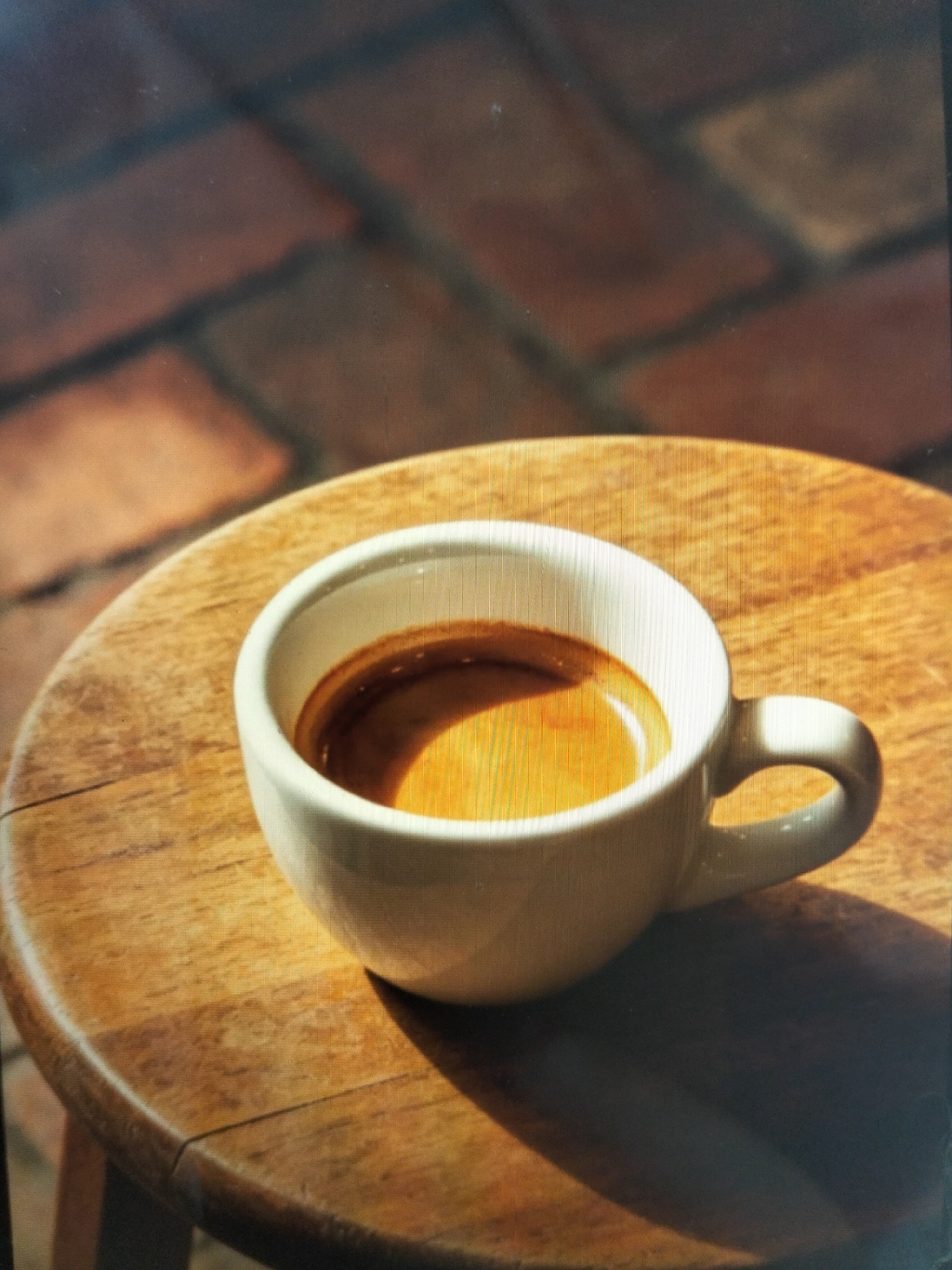 A close-up, high-angle shot features a white espresso cup filled with dark coffee, resting on a round wooden table. The coffee has a visible crema layer. The table surface shows wood grain and appears to be outdoors, with sunlight casting a shadow from the cup. In the background, out of focus, is a patterned surface resembling brickwork. The scene is set in the city of Payagaboh, Indonesia.