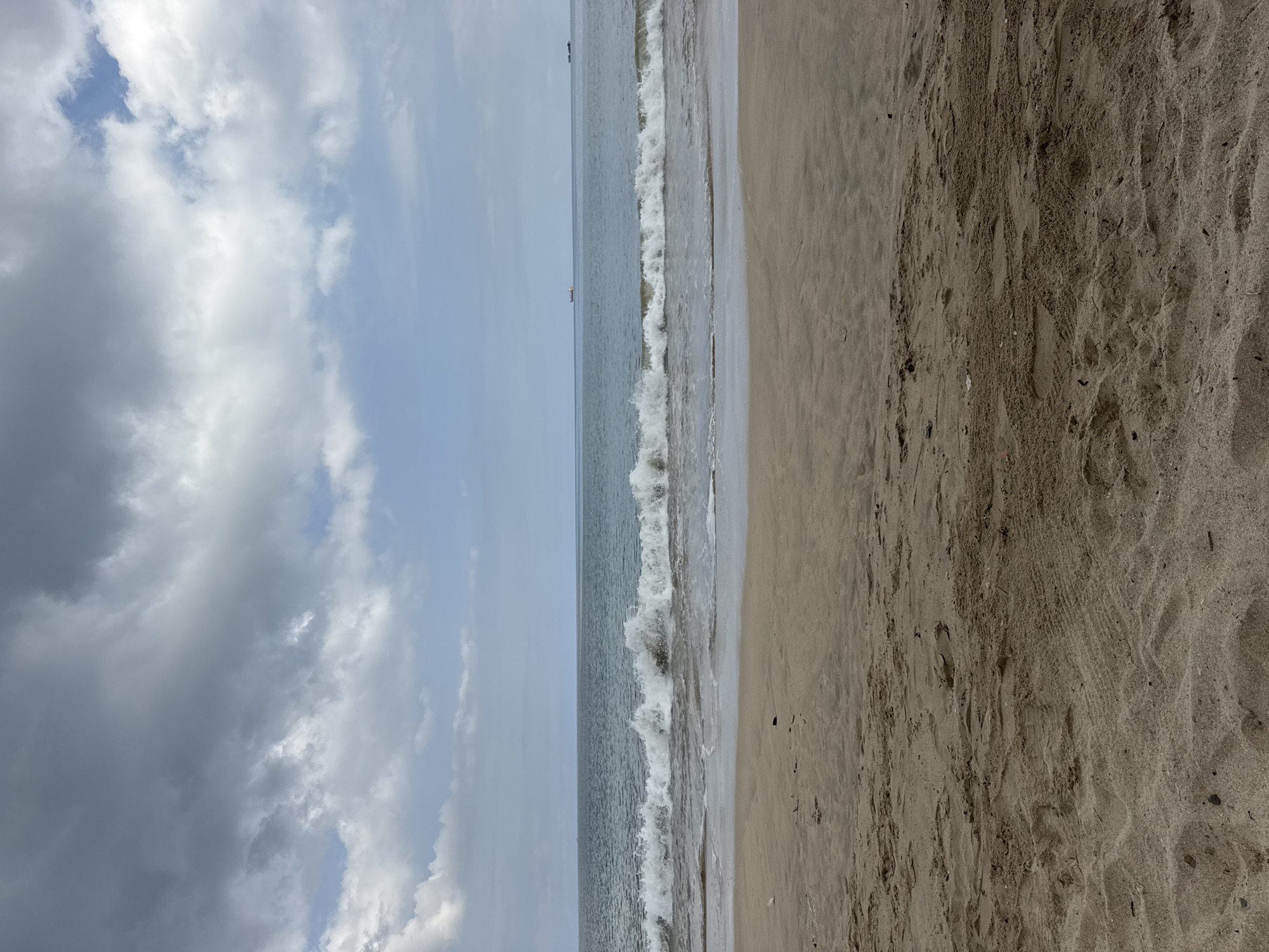 The image depicts a beach scene in Kuta Selatan, Indonesia. The composition is dominated by a sandy beach on the right, transitioning into a shoreline with small waves breaking. Beyond the shoreline is the ocean under a partially cloudy sky on the left. There are no visible people or distinct objects besides the sand, water, and clouds. Two tiny objects that seem like boats are visible off-shore. The sand is coarse and uneven, indicating natural terrain. The waves are small and foamy, suggesting mild ocean conditions. The sky consists of both light blue areas and darker, overcast clouds, indicating a partly cloudy day.