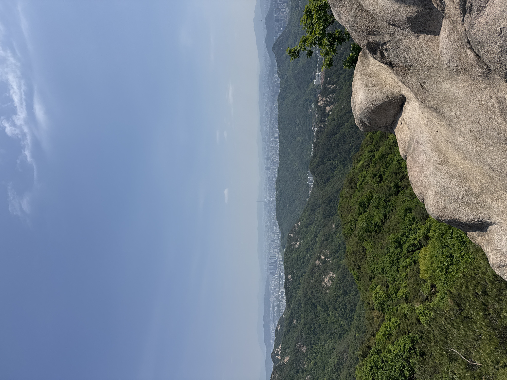 The image shows a view overlooking Seoul, South Korea, from a high vantage point, likely a mountain. In the foreground to the right is a large, light gray rock formation with some green foliage visible around it. A dense, green forest covers the lower slopes of the mountain. A cityscape is visible in the distance, with multiple buildings and structures forming the urban landscape. The sky above the city is a pale blue, hazy, with light cloud cover on the left. The image appears to be taken during the daytime in a clear, sunny, though hazy day.