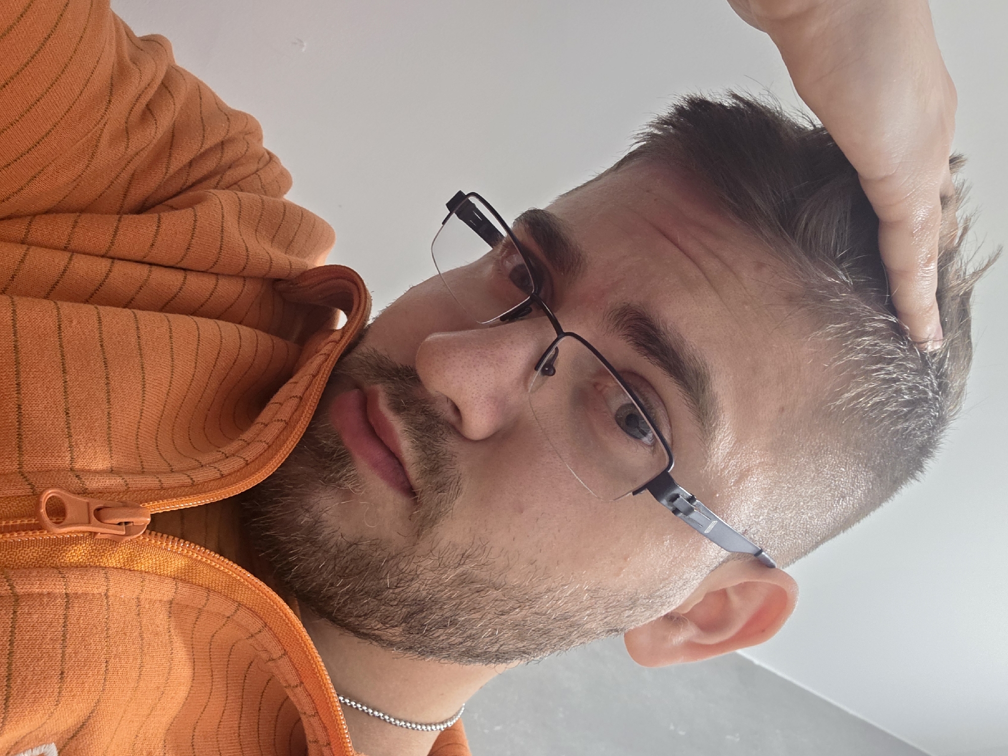 A close-up, selfie-style photograph presents a man with fair skin, short dark hair with hints of grey, and a beard. He is wearing glasses with thin, rectangular black frames. He has light-colored eyes and a slightly furrowed brow. His lips are pink and full. He is wearing a light orange zip-up hoodie. A silver chain necklace is visible around his neck. His right hand is resting on the top of his head, fingers touching his hair. The background is a plain white surface. No specific location in Plovdiv, Bulgaria, is discernible from the image.