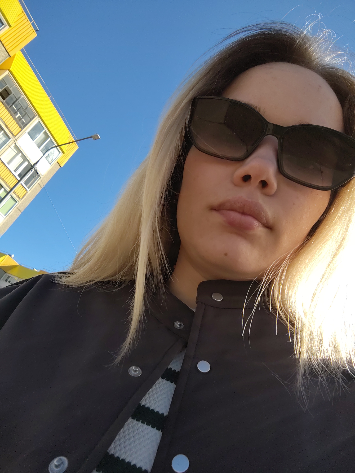 A medium-close-up, low-angle shot captures a woman outdoors, appearing to be taking a selfie. She is fair-skinned with long, blonde hair that cascades over her shoulders and is brightly illuminated by sunlight. She is wearing large, dark sunglasses that obscure her eyes, and her lips are slightly parted. Her expression is neutral. She is dressed in a dark, possibly black or navy, collared jacket with silver snap buttons visible on the front and shoulders. Beneath the jacket, a garment with light blue and black horizontal stripes is visible on her left side.
The background features a clear, bright blue sky dominating the upper left portion of the image. To the far left, a tall building with prominent yellow panels on its facade and multiple windows is visible, stretching upwards from the bottom left corner. A street light or lamppost extends from the building into the blue sky. The lighting indicates a sunny day. No text is visible in the image. The scene is set in what appears to be an urban or suburban environment in Марковский, Russia.