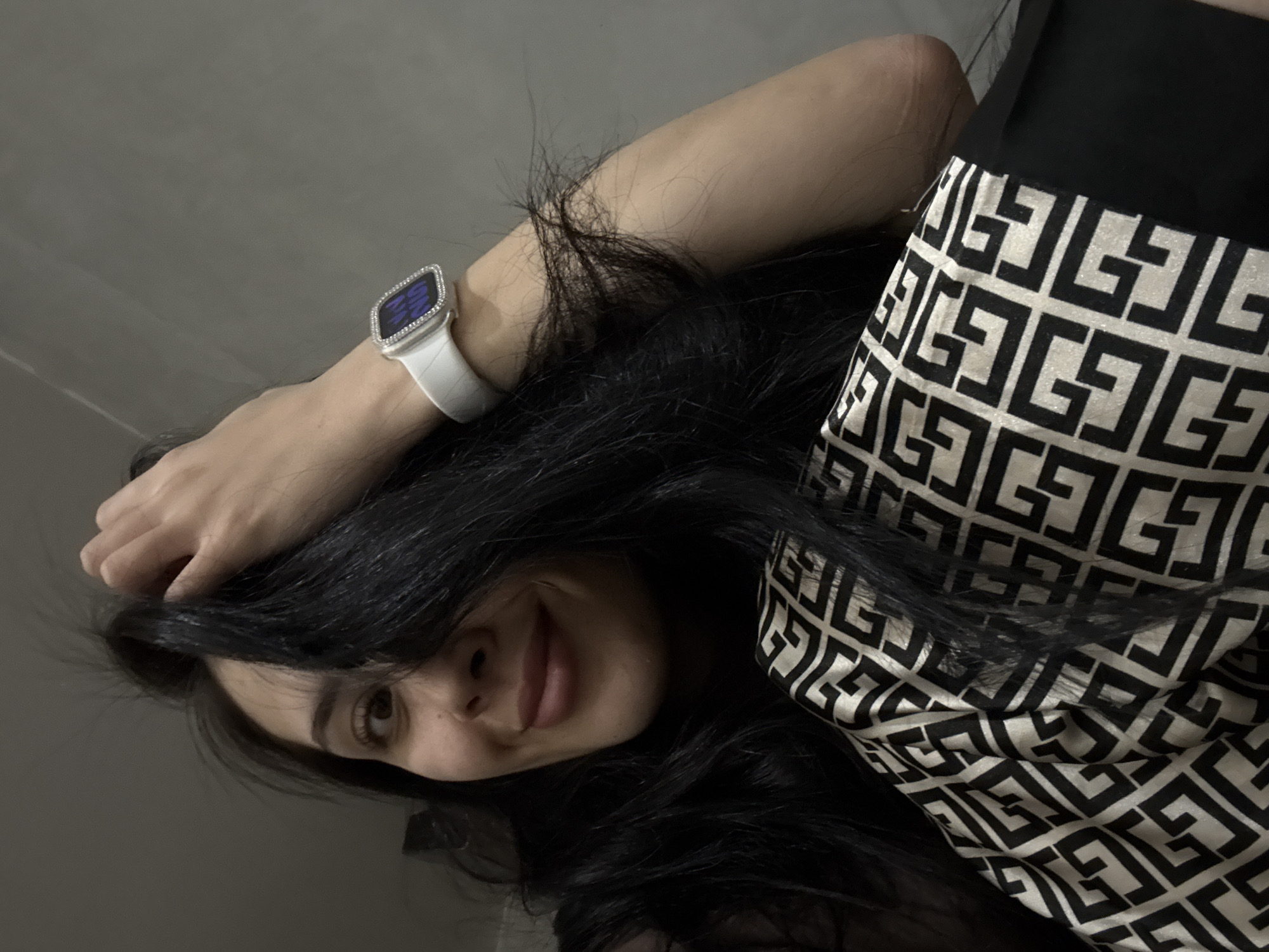 A woman with dark hair is lying down, looking at the camera with a slight smile. She is wearing a white watch with a silver bezel. Her clothing appears to be a skirt or dress with a geometric black and white pattern. The background is a plain, light-colored wall. There is no indication of Votkinsk, Russia or any specific location.