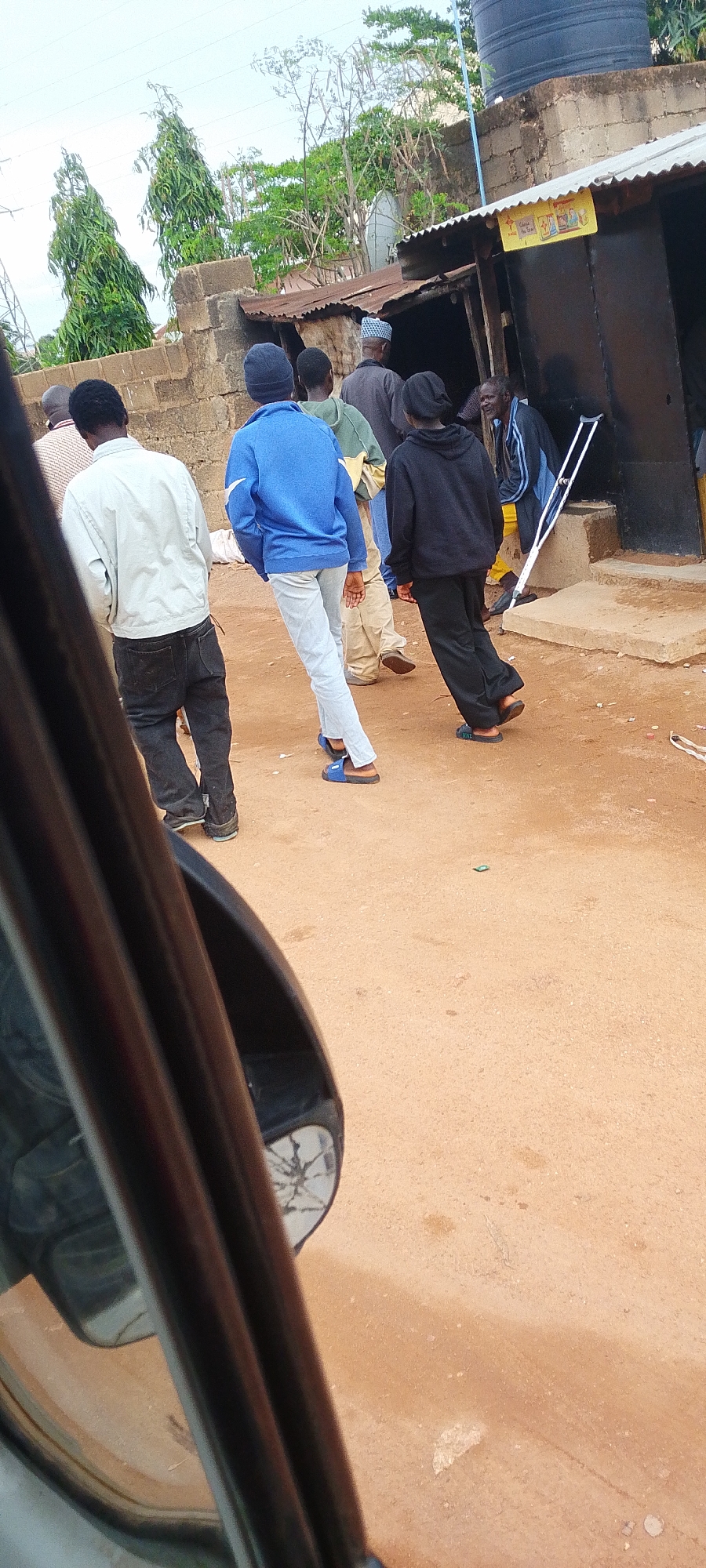 The image depicts a street scene in Jos, Nigeria, with several individuals in the foreground and middle ground. A person wearing a light-colored jacket and dark pants is walking away from the camera. To their left, another person in a blue hoodie and light-colored pants is also walking away. Behind them, several other individuals are visible, some with their backs to the camera, walking towards a building. The building appears to be a small shop or structure with a corrugated metal roof. A person is leaning on a crutch near the entrance of the building. The ground is unpaved and appears to be dirt or sand. In the upper right portion of the image, a large black water tank is visible on top of a building. Trees and vegetation are present in the background. The overall scene suggests a casual gathering or movement of people on a street in a Nigerian urban or semi-urban setting.