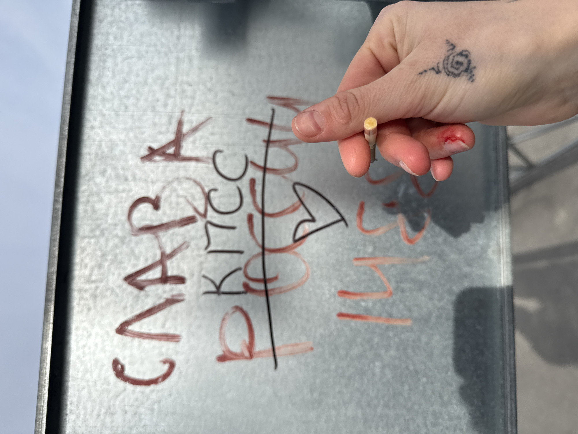 The image depicts a hand holding a cigarette butt in front of a metal surface with graffiti. The graffiti includes the word "САЛА" (SALA) and the numbers "143". There are red marks near the fingernail on the index finger of the hand. The hand also has a tattoo. The location cannot be determined based solely on the image.
