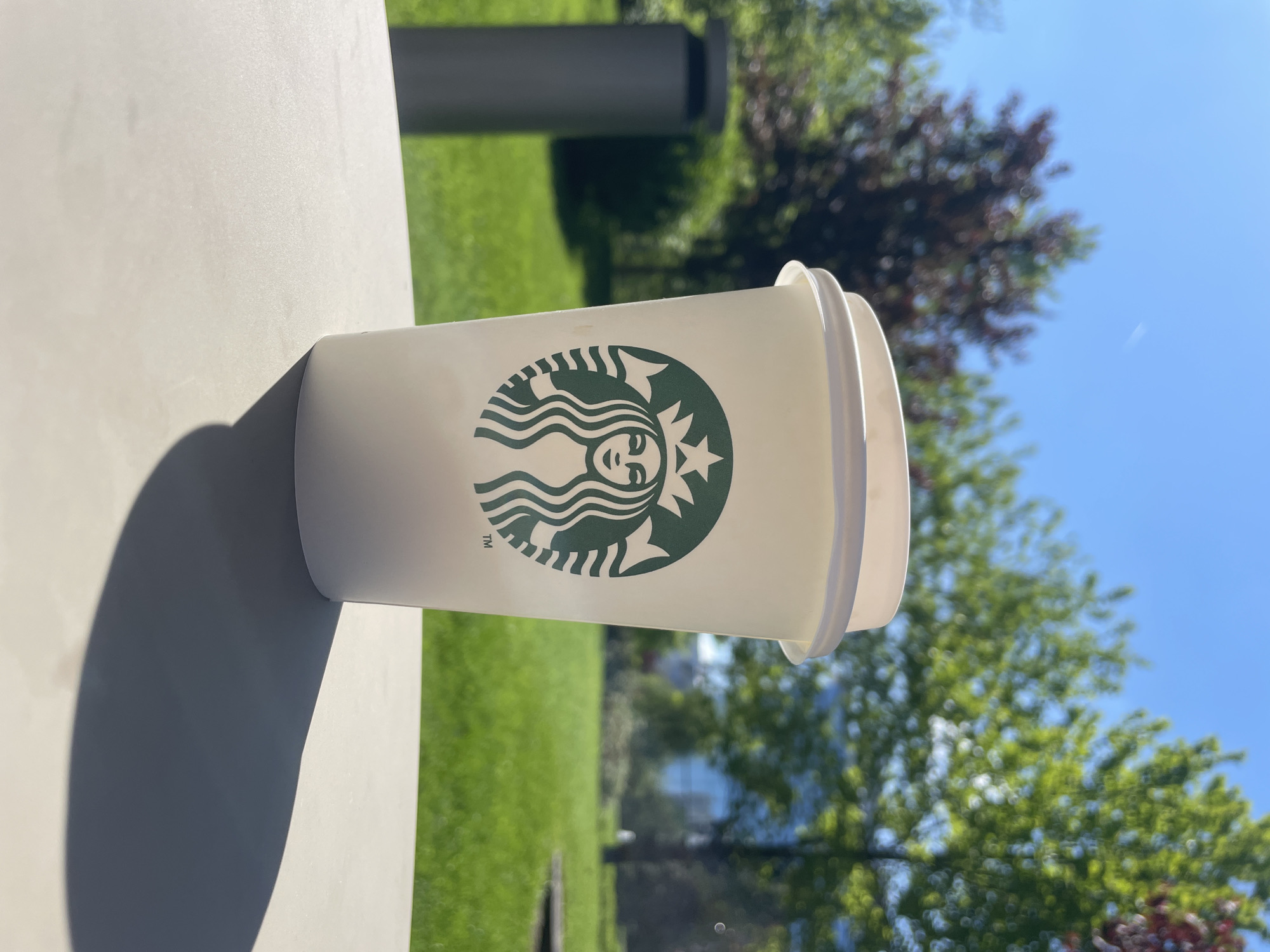 A Starbucks coffee cup with a lid is on a gray surface. A shadow is cast on the surface. The background shows a grassy area, trees, and a blue sky. It appears to be outdoors. It's difficult to determine the exact location in Sofia, Bulgaria, without more information.