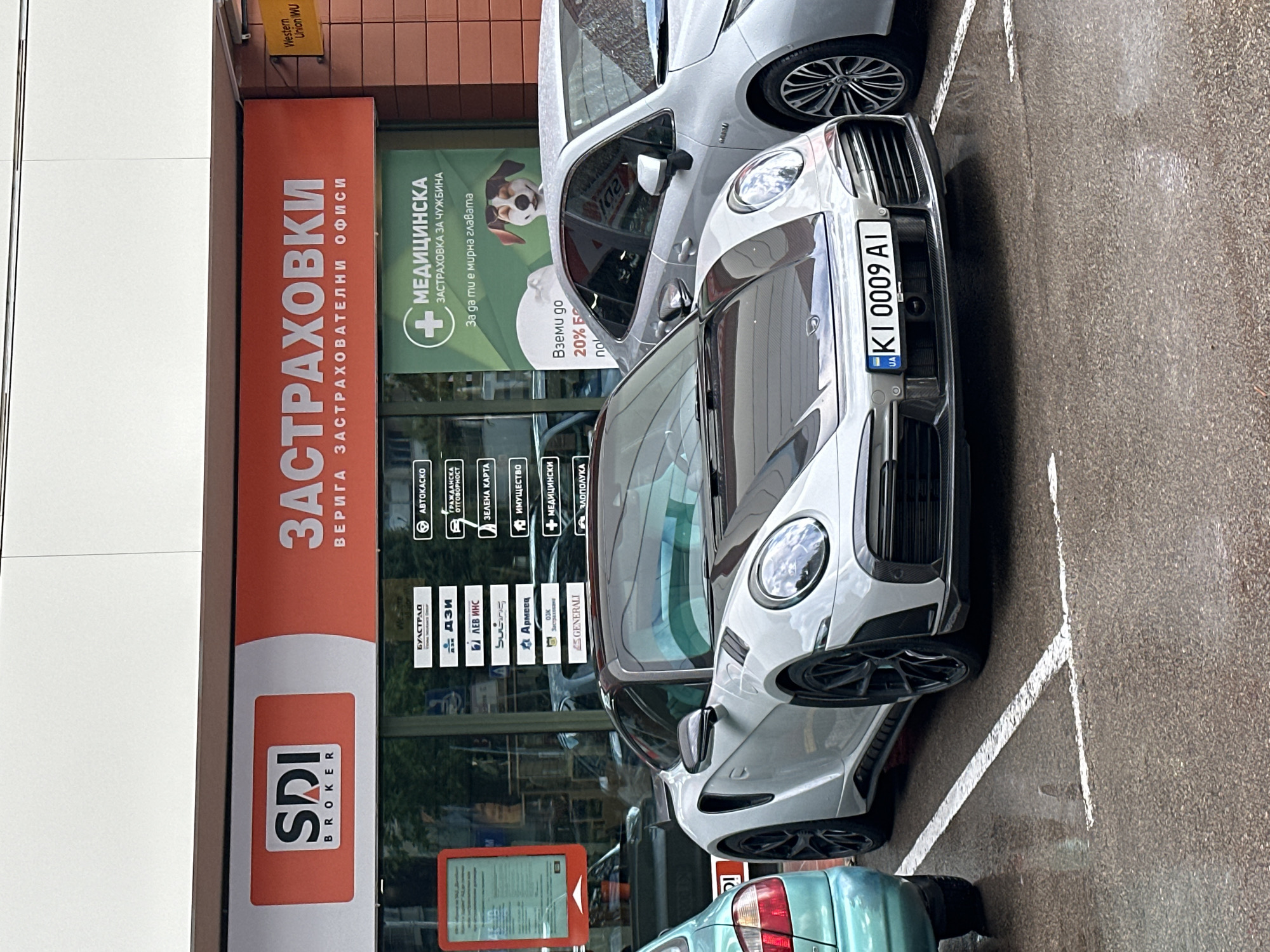 The image shows a parking area outside an SDI Broker office in Varna, Bulgaria, specializing in insurance. The office has a large red sign with "ЗАСТРАХОВКИ" (Insurance) written in Cyrillic script, and lists different types of insurance they offer, including автокаско (vehicle insurance), гражданска отговорност (civil liability), and medical insurance. A green sign advertises medical insurance for foreigners ("МЕДИЦИНСКА ЗАСТРАХОВКА ЗА ЧУЖБИНА") featuring an image of a dog. In front of the office are several parked cars, including a silver sports car with the license plate KI 0009 AI. The ground appears wet, possibly from rain. A Western Union sign is visible in the background.