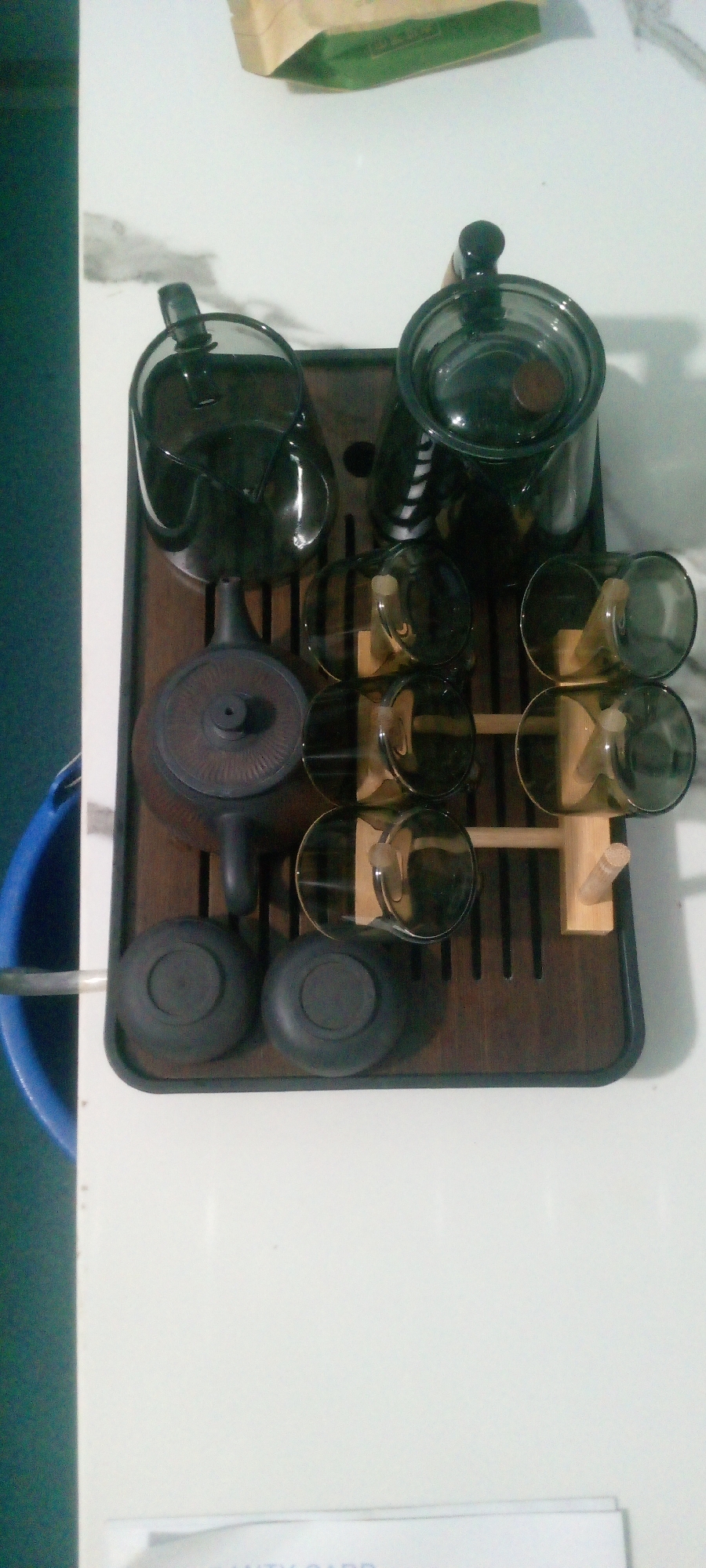 The image shows a tea set on a dark wooden tray. The tea set includes a teapot, a pitcher with a lid, and several glasses. The pitcher is large and clear with a black lid and handle. The teapot is smaller and made of a dark material with a spout and lid. There are eight glasses, arranged in two rows of four, resting on wooden stands. The glasses are clear and appear to be filled with a dark liquid. Two round, dark lids or coasters are placed at the bottom left of the tray. The tray itself has a slatted design. In the background, a green and beige bag is visible, and a blue bucket can be seen to the left of the tray. The scene is indoors and appears to be a tabletop. There is no indication of the city of Indo, Nigeria.