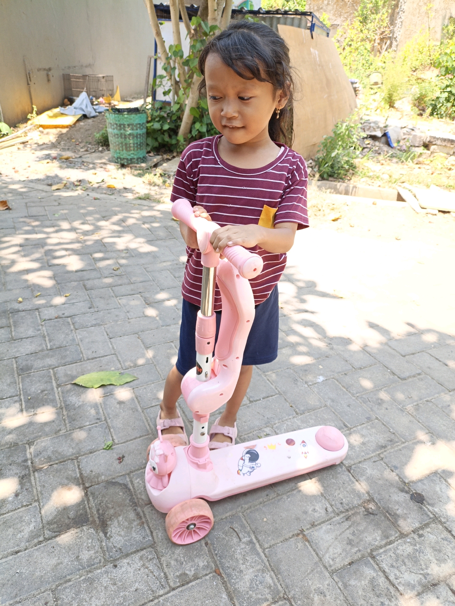 A young girl stands on a paved outdoor area in Cikal, Indonesia, holding onto a pink and white scooter. She is wearing a maroon and white striped t-shirt and dark blue shorts. Her feet are clad in pink sandals. The scooter is a three-wheeled model with the front two wheels slightly elevated. The scooter deck is decorated with stickers, including an astronaut. The ground is made of interlocking gray pavers, with scattered dry leaves. In the background, there is a wall, some greenery, and a green trash can. The lighting suggests it is daytime, with sunlight casting shadows.