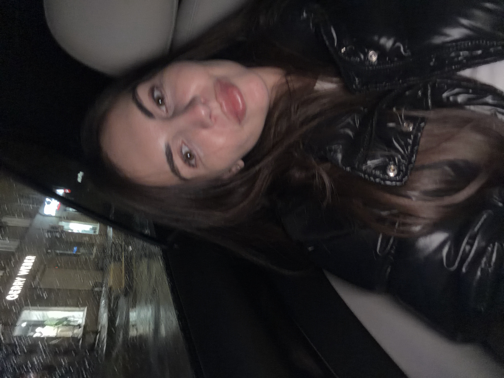 A woman is in the backseat of a car. She is wearing a black, shiny puffer jacket and has long, brown hair. The car appears to be stopped or moving slowly. Through the car window, a storefront with the name "Gerry Weber" is visible, along with reflections of lights and signs. It is raining outside. Based on the information, it can be inferred that the location is Krasnoyarsk, Russia.