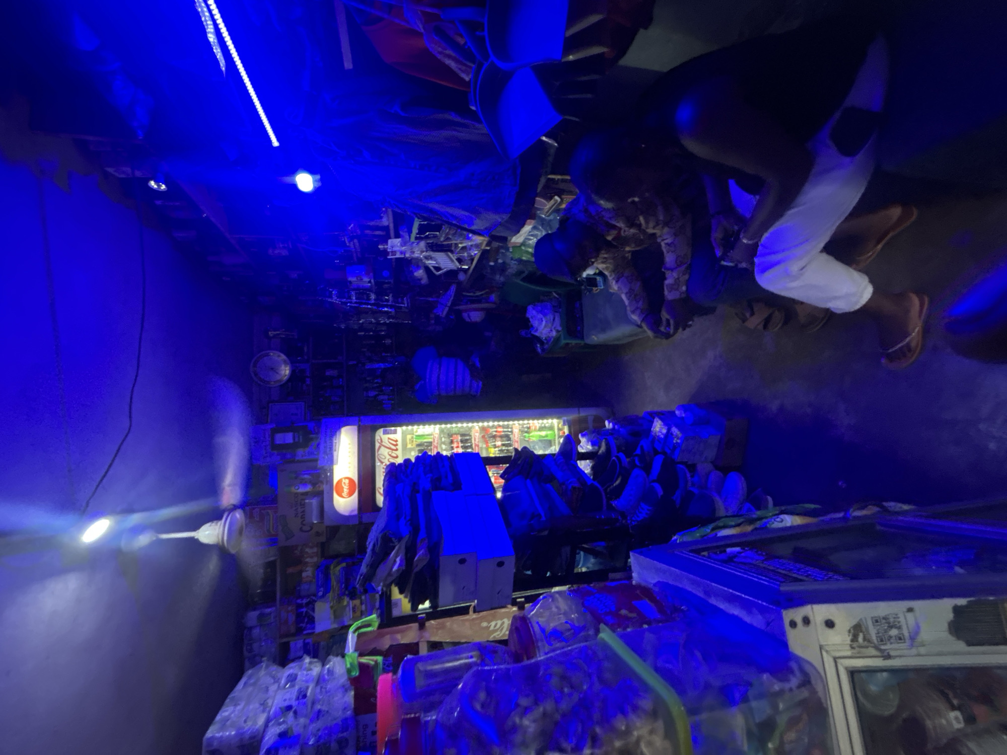 The scene depicts the interior of a small, cluttered shop in Jos, Nigeria. The primary lighting is dominated by a bright blue hue, likely from LED strips, which casts an unusual color balance throughout the space.
On the right, two individuals are present. One is kneeling, possibly engaging in a transaction. Both wear casual clothing, with one person sporting white trousers and sandals.
Shelves filled with various merchandise occupy the space behind the seated figures. A Coca-Cola vending machine and a spinning ceiling fan are also visible, suggesting that the shop is selling beverages and necessities. Rows of bottled water or similar products are stacked near the lower part of the frame.
The shop appears densely packed with goods, creating a cramped atmosphere. Details on individual items on the shelves are difficult to discern due to the lighting conditions and image orientation.