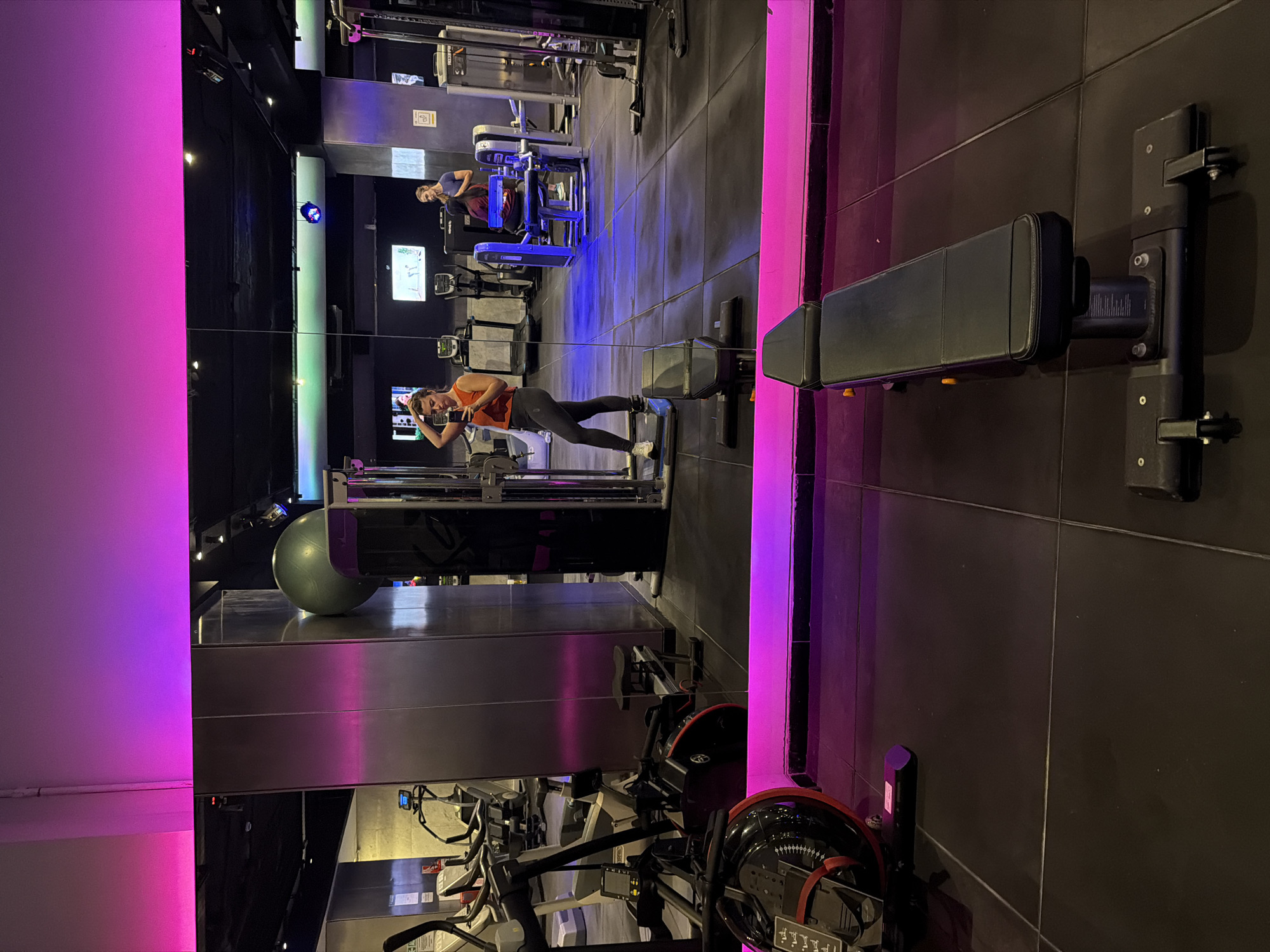 A brightly lit gym interior. A woman dressed in a red top and black leggings is seen taking a photo of herself in a mirror while leaning against gym equipment. In the mirror's reflection, another person is visible seated on a leg extension machine. Various gym machines and equipment are also present in the scene. Given that it's in Buenos Aires, Argentina, the environment has a modern, upscale feel.