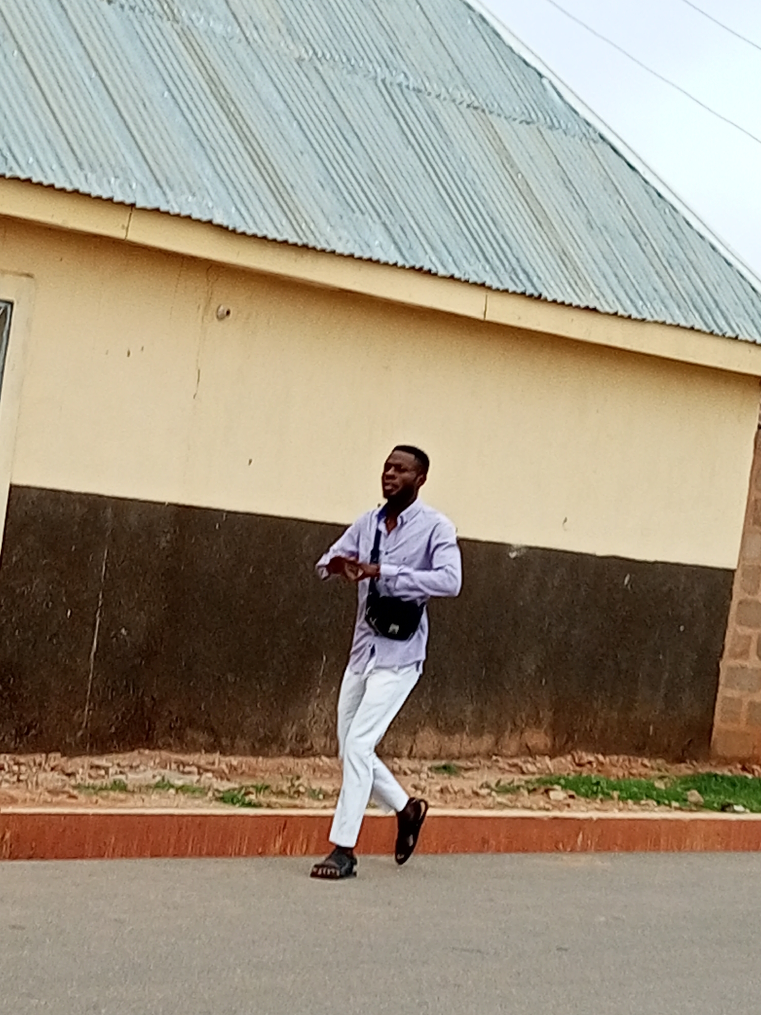 A man with dark skin and short dark hair is walking on a paved surface. He is wearing a light purple long-sleeved collared shirt, white pants, and black sandals. A black crossbody bag is worn over his right shoulder and across his chest. He is looking to his left with his right arm extended forward and his left arm bent at the elbow. He appears to be in motion. Behind him, there is a building with a light yellow upper half and a dark brown lower half. The building has a corrugated metal roof. A strip of grass and dirt separates the building from the paved walkway in front of it. The walkway has a reddish-brown curb. The sky above is overcast.