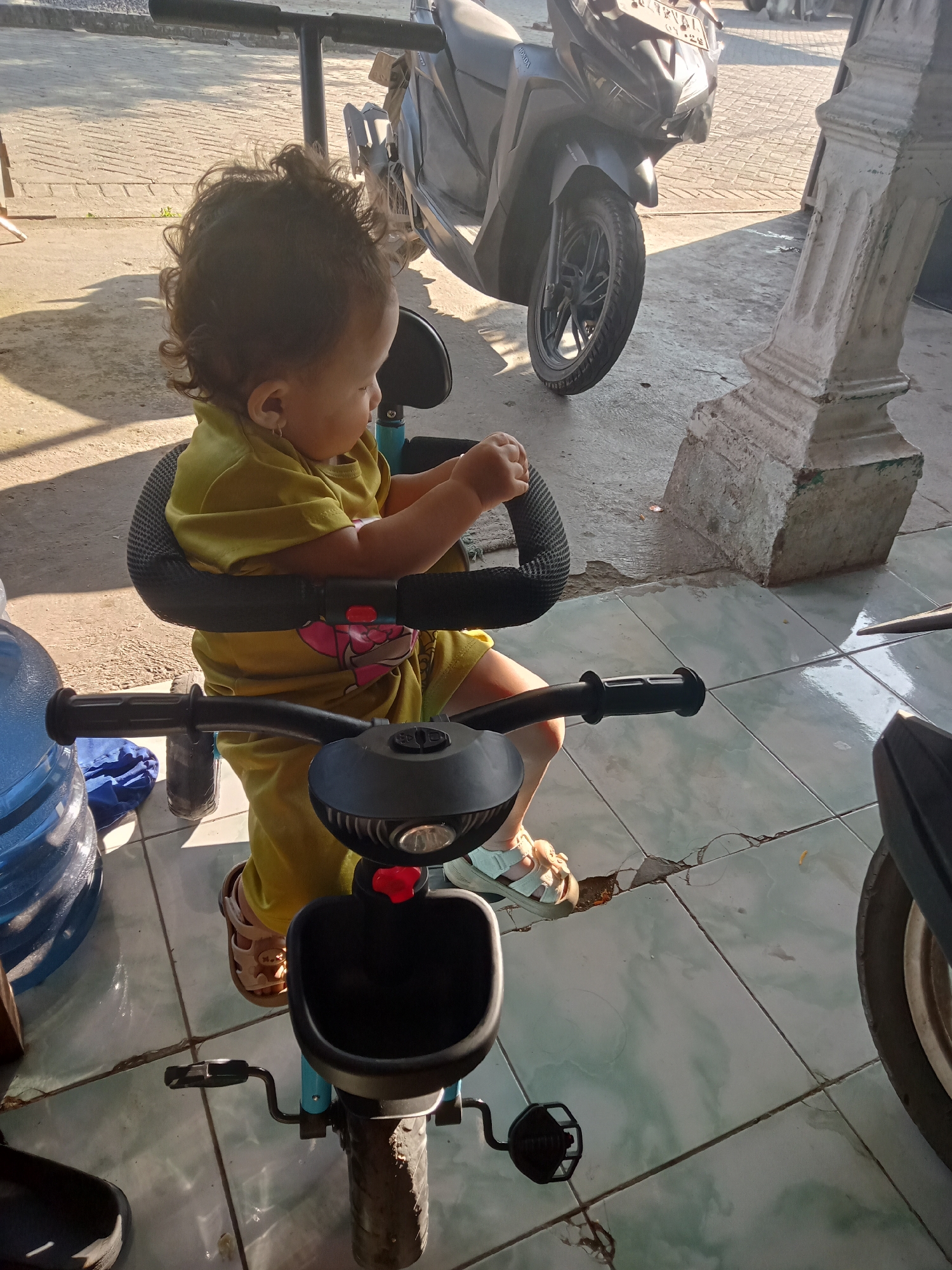 In Bungur, Indonesia, a young child, wearing a yellow outfit and sandals, sits on a blue and black tricycle. The child is positioned on a tiled floor in what appears to be an outdoor or semi-outdoor space. Adjacent to the tricycle and child is a large, blue water jug. In the background, there is a black motorcycle parked on a paved surface. A white architectural pillar stands nearby. The environment suggests a residential or commercial area within Bungur.