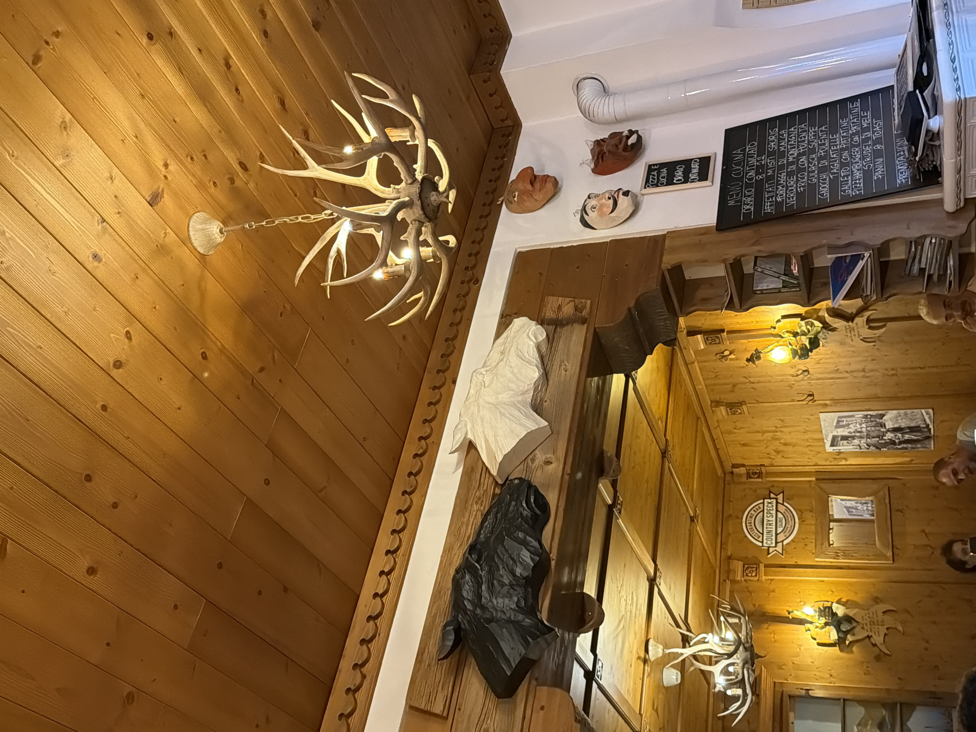 The image depicts the interior of a restaurant with a rustic, wooden decor, possibly located in Sauris di Sopra / Plozn / Sauris Disore, Italy, given the "COUNTRY SPECK SAURIS" sign visible. The ceiling is paneled with light-colored wood. A prominent antler chandelier hangs from the ceiling, featuring multiple light sources. On the wall, several decorative items are displayed, including carved wooden horse heads (one black, one white) and three decorative masks. To the right, a white ventilation pipe extends upwards. A black chalkboard menu is mounted on the wall below the masks. The menu lists "PIZZA E CUCINA" and other food items in Italian. The overall ambiance suggests a traditional mountain restaurant. In the lower portion of the image, a small section of a fireplace or tiled structure is visible, along with several people seated, partially visible. The lighting is warm and ambient, contributing to the cozy atmosphere.