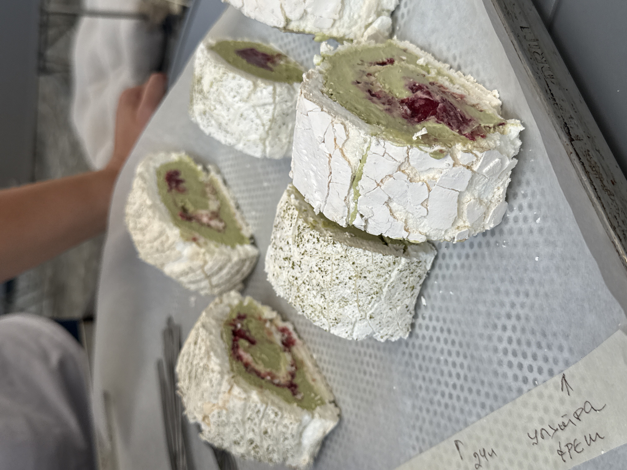 A close-up, slightly overhead shot captures several portions of a matcha roulade dessert resting on a white, textured paper lining inside a metallic baking tray. The roulades, light in color and coated with what appears to be meringue, are sliced to reveal a green filling, likely matcha cream, and interspersed with red pieces, possibly berries or jam. Some of the roulades are dusted with a fine green powder, which could be more matcha.
In the background, a person's torso and arm are partially visible, suggesting they are involved in preparing or arranging the dessert. The environment appears to be a kitchen or a food preparation area, indicated by the baking tray and the overall setting.
The lower left corner of the image shows a piece of white tape with handwritten text in a non-Latin script. This tape may be a label or instruction related to the dessert. The baking tray has visible markings on its right edge, including the word "DRIN."
The scene is well-lit, with the focus on the dessert. No specific landmarks or elements definitively place the location in Vracar Urban Municipality, Serbia, beyond the user's request to include it.