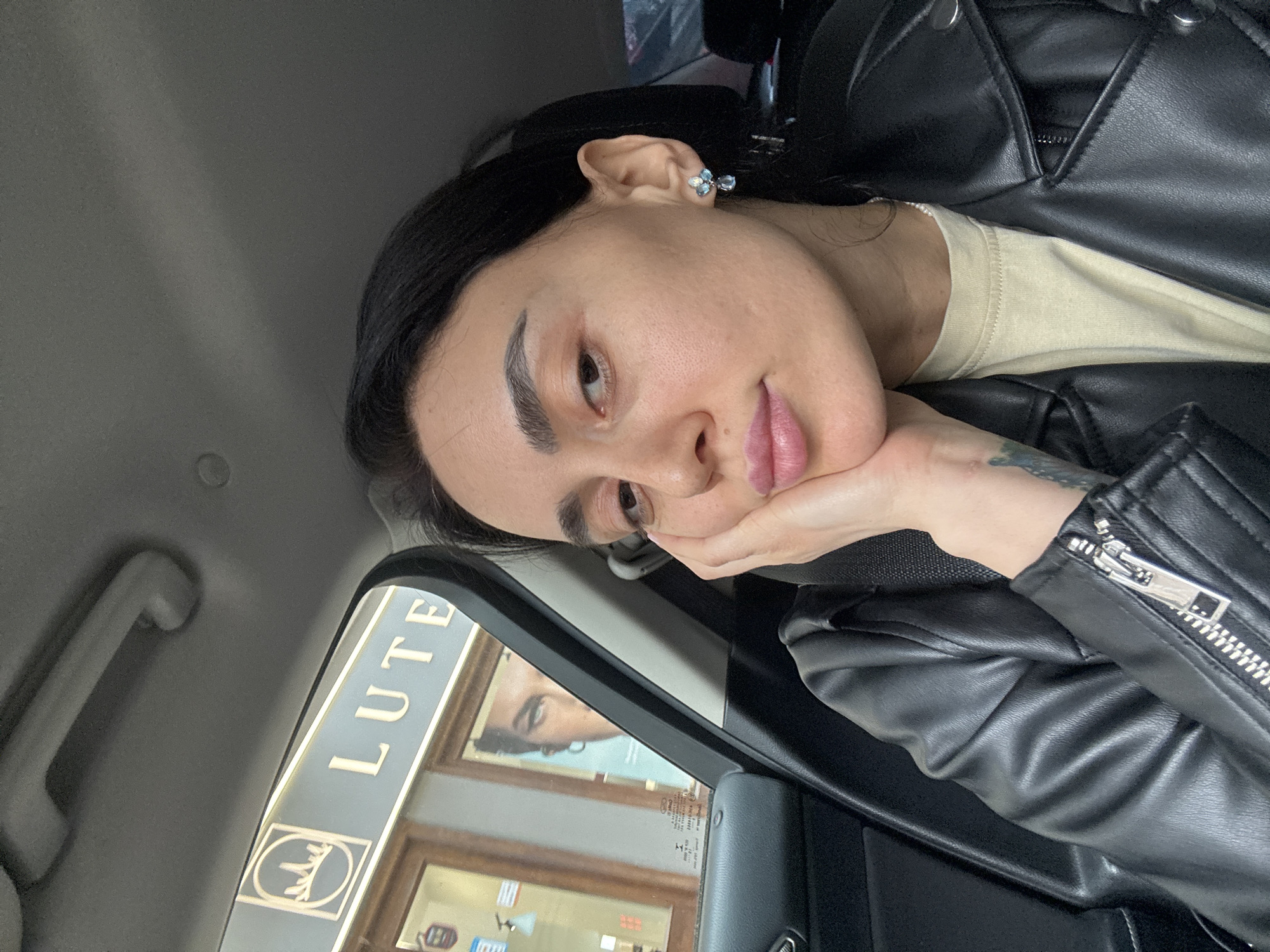 A female subject with dark hair and light skin is positioned in the passenger seat of a vehicle, taking a selfie. She wears a black leather jacket over a light beige t-shirt. Her left hand rests against her left cheek, with fingers extended towards her chin, and her lips are slightly pursed. She has blue and silver multi-stone earrings in her right ear, and a faint tattoo is visible on her left forearm. The vehicle's interior is dark grey, with a grab handle visible on the ceiling and a seatbelt across her chest.
Through the vehicle's window on the left, a storefront is visible. A sign with the word "LUTE" in large white letters on a light blue background is present, along with a stylized white logo resembling a cityscape or crown within a square. This identifies the location as Lute Perfume House, located at 3 Shota Rustaveli Avenue in Tbilisi, Georgia. Inside the storefront window, a large advertisement poster featuring a close-up of a person's eyes and upper face is visible. A small red fire extinguisher symbol is also discernable inside the store.