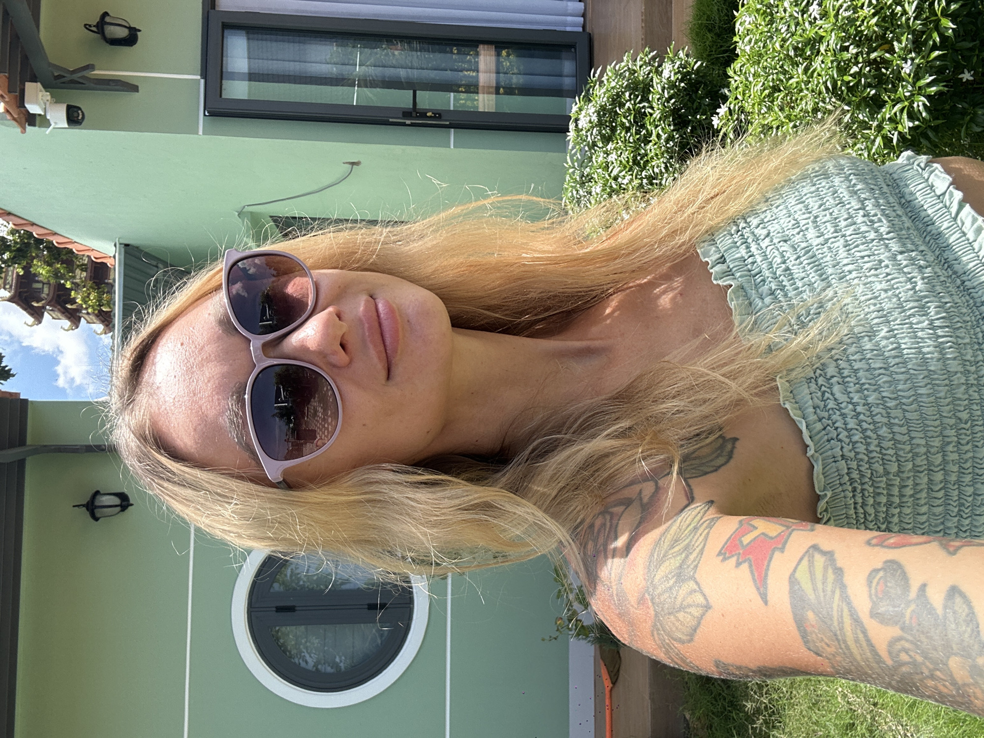 A fair-skinned woman with long, blonde hair, wearing light purple sunglasses and a light green ruched strapless top, is depicted in an outdoor portrait. Tattoos are visible on her right arm and shoulder. The background features a light green building with a dark brown roof trim, which includes a circular window with a white frame on the left and a black-framed glass door on the right. Black outdoor lanterns are mounted on the building's facade, along with a white security camera positioned above the door. Green grass is visible in the lower-left, and bright green bushes with small white flowers are in the lower-right. In the distant background, behind the building and against a blue sky with white clouds, a multi-tiered traditional Vietnamese pagoda structure with ornate, curved roofs is visible. This pagoda structure identifies the location as being in the vicinity of the Linh Sơn Trường Thọ Pagoda on Ta Cu Mountain, near Mũi Né, Vietnam.