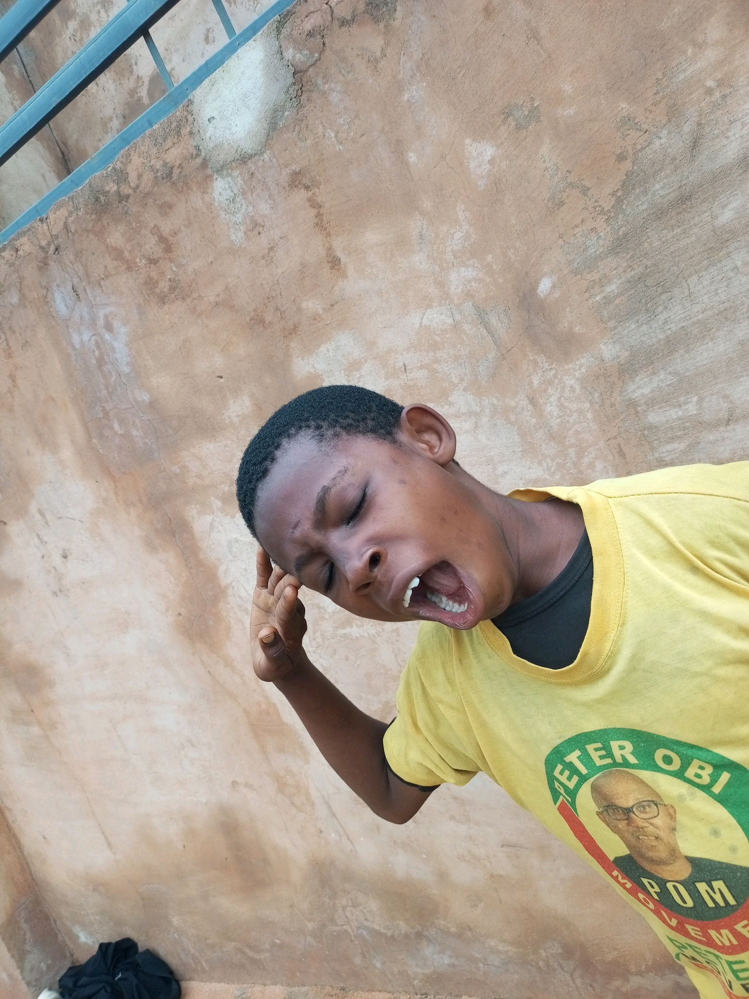 A young Black boy is pictured from the chest up, leaning his head back and to the right with his eyes closed. His mouth is wide open, revealing his teeth. His right hand is raised and his index finger is touching his forehead. He is wearing a bright yellow t-shirt with a black collar. A circular graphic is printed on the front of the t-shirt, featuring a portrait of a man wearing glasses, with the words "PETER OBI MOVEMENT" around the edge.
The background is a rough, textured wall, appearing to be made of a sandy, concrete-like material. The wall has patches of lighter and darker discoloration, and some visible cracks. In the upper left corner of the image, a portion of a blue metal railing or bar is visible, running horizontally. Below the boy's t-shirt, on the lower left, a crumpled piece of dark clothing is visible on the ground. The lighting suggests it is daytime, with harsh shadows cast by the railing. The overall scene appears to be outdoors, possibly in an urban or semi-urban environment, given the wall and railing. The specific location of Tempure, Nigeria is not identifiable from the visual information in the image itself.