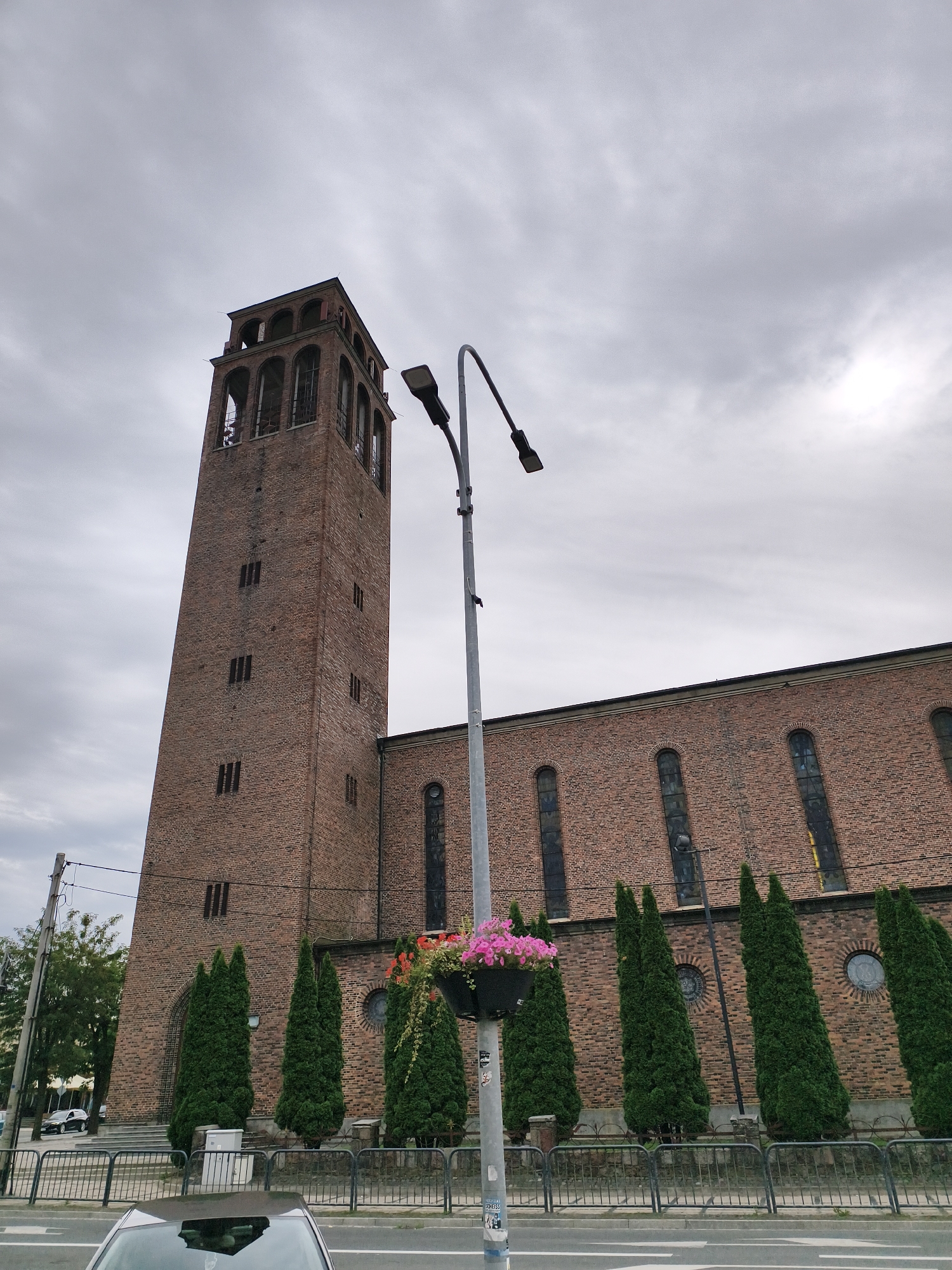 The image depicts the Church of Saint Stanislaus the Bishop and Martyr in Tczew, Poland. The imposing brick structure is characterized by its tall, slender bell tower adorned with multiple arched openings. A long, red brick nave extends to the right, featuring a series of arched windows. In the foreground, a street is visible with a railing separating it from the church grounds. Several tall, conical evergreen trees are planted along the fence. A modern lamppost with two lights stands prominently in the middle ground, decorated with a planter box filled with pink flowers. The sky is overcast and gray, suggesting a cloudy day.