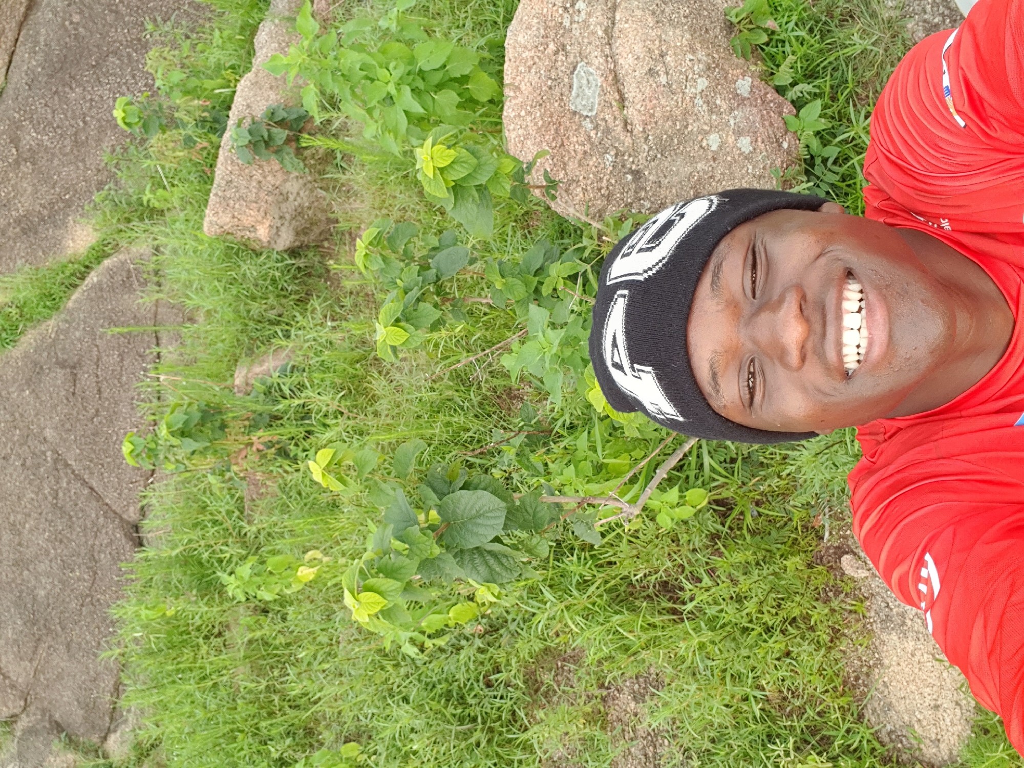 A young person is captured in a selfie, their face filling the upper right portion of the frame. They are wearing a black beanie with white lettering and a bright red long-sleeved shirt. The individual is smiling broadly, showcasing their teeth and exuding a joyful emotion. The photo appears to be taken outdoors, with a background of lush green grass and several large, weathered rocks. Some small green plants are interspersed with the grass. The lighting suggests it is daytime. The overall scene conveys a relaxed and happy moment in a natural, possibly rocky outdoor environment, consistent with the location context of Jos, Nigeria. No discernible text is present on the beanie or clothing besides decorative patterns.