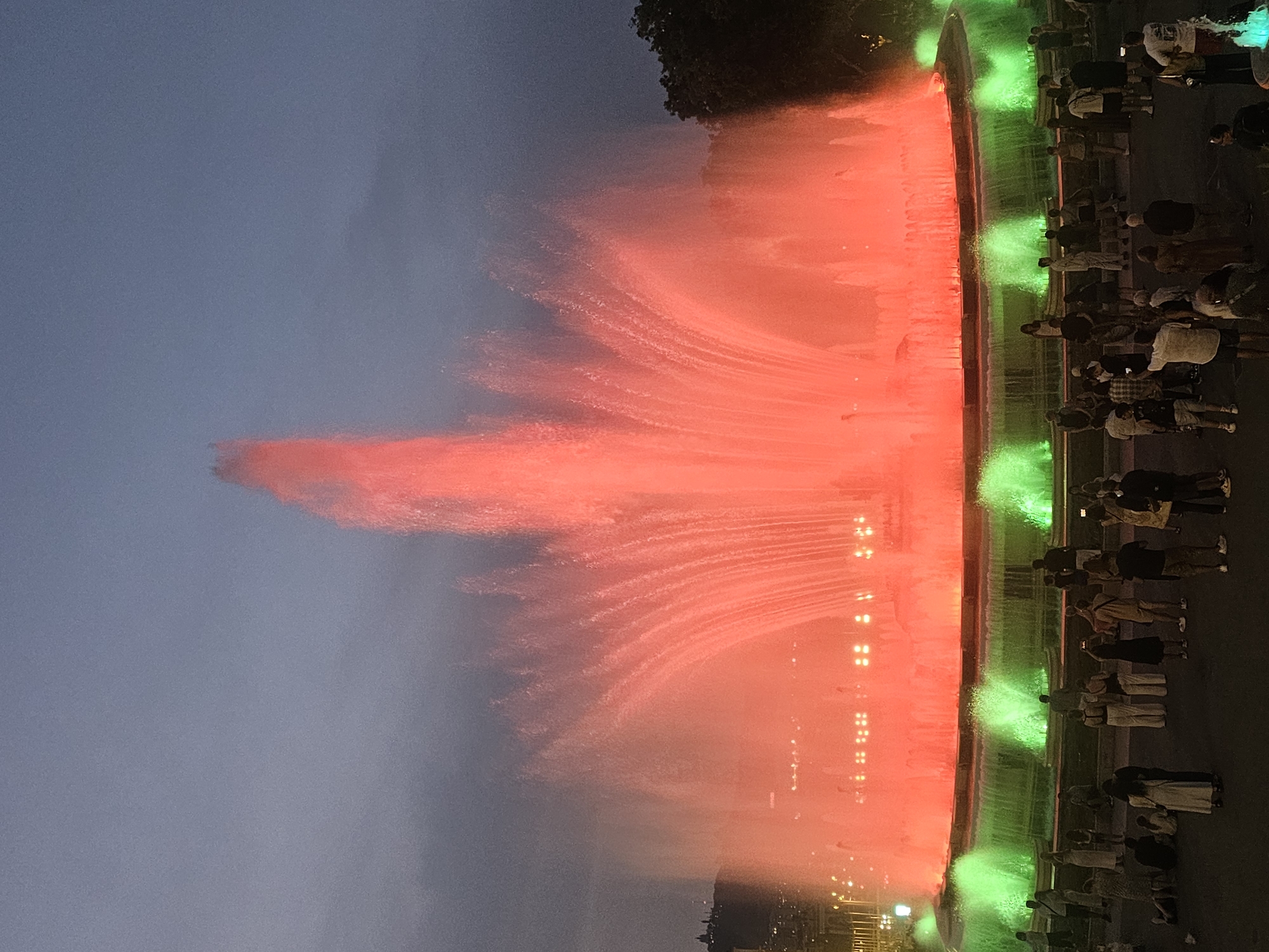 This is a photograph of the Magic Fountain of Montjuïc in Barcelona, Spain. The fountain is illuminated with red and green lights, creating a vibrant display against the twilight sky. Several people are gathered around the fountain, observing the spectacle. The fountain is the central focus of the image, with its water jets reaching high into the air. The surrounding area is dark, with some distant lights visible, suggesting an urban setting. The overall impression is one of a public event or attraction drawing a crowd.