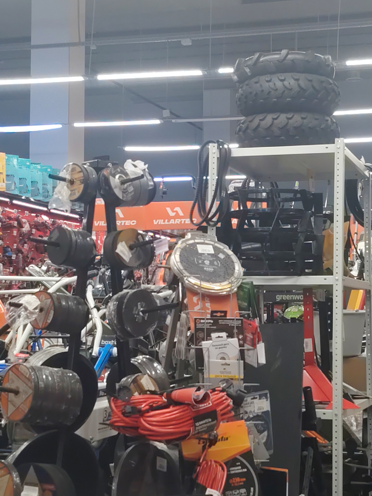 The image shows a retail store environment, likely a hardware or home improvement store. In the foreground, there are several racks of goods. One rack displays stacks of heavy black tires, possibly for agricultural or off-road vehicles. Another rack holds weight plates for barbells, made of dark gray metal. There are also spools of red electrical cord. In the background, more shelving units are visible, filled with various products, including boxes and tools. The store appears well-lit with overhead fluorescent lighting. The overall scene depicts a retail display of tools and equipment.