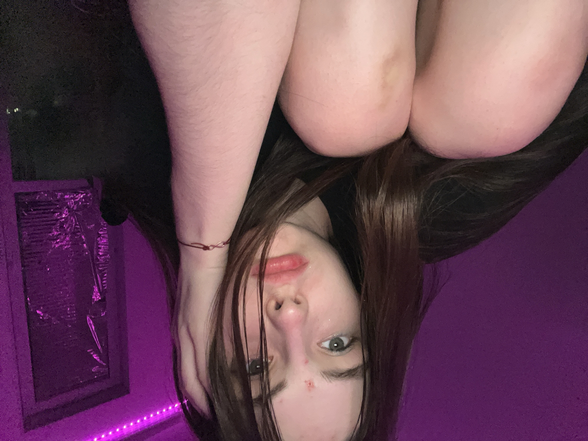 A young woman is visible upside down in the frame, resting her knees on her head. She has long brown hair with some strands falling across her face. She is wearing a black shirt. The background includes a window with reflective material partially covering it, illuminated by purple LED lights. One wall is a solid purple color. A bracelet is visible on her wrist.