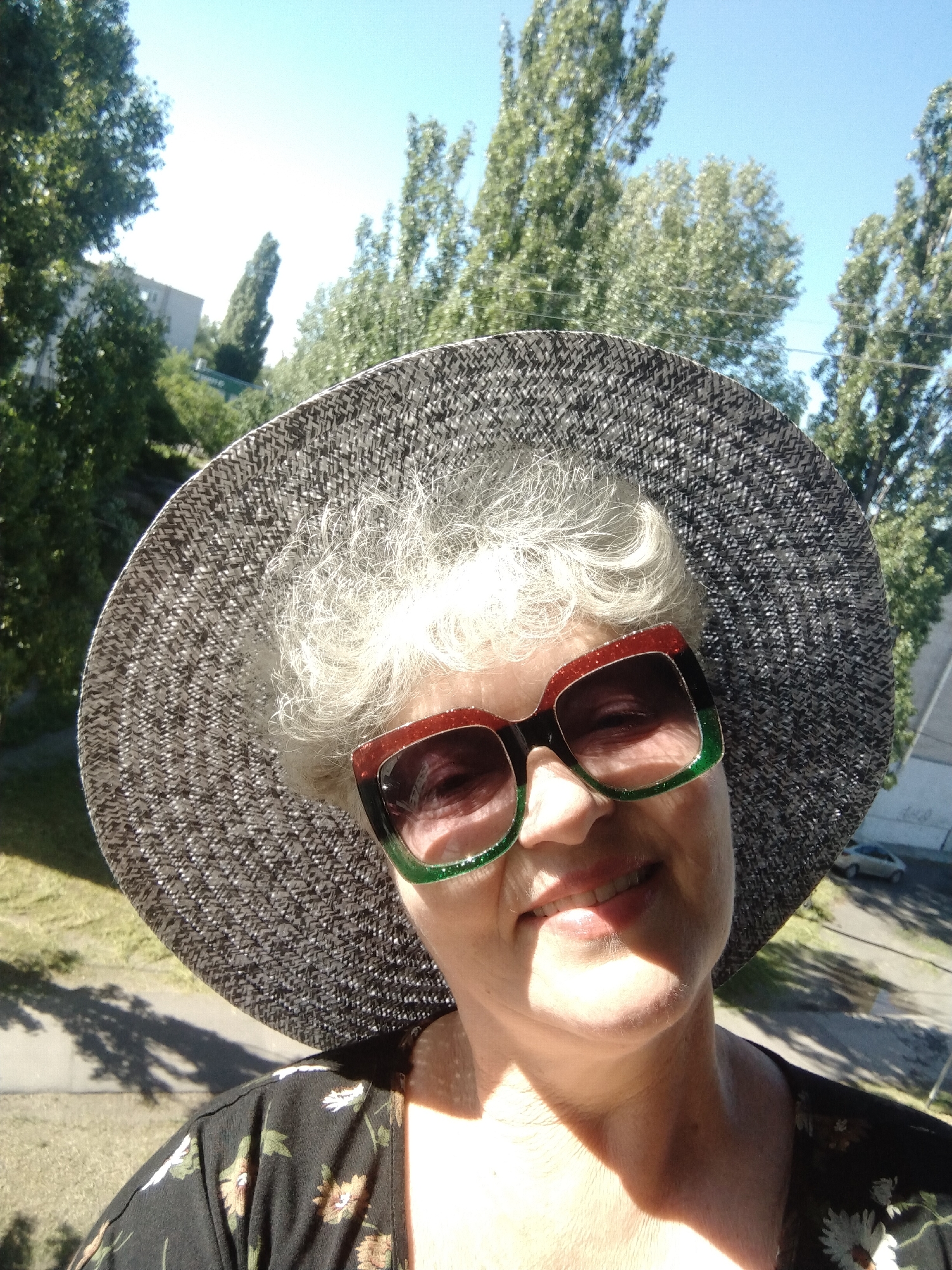 A woman wearing a large sun hat and sunglasses is smiling in a selfie. She is wearing a black floral patterned shirt. The background shows trees and a building. A road with a car on it is visible. The location is Horishni Plavni, Ukraine.
