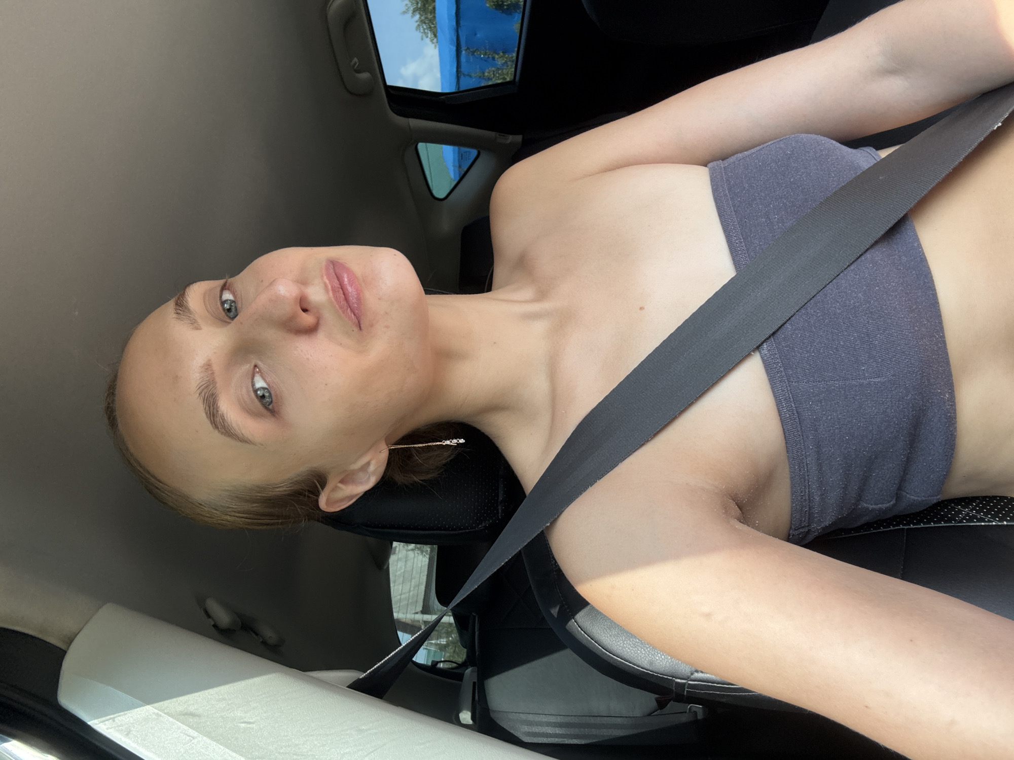 A woman is in the driver's seat of a car, looking at the camera. The woman has blonde hair pulled back and is wearing a grey strapless top and a black seatbelt. She has on long, dangly earrings. The interior of the car is visible, with black leather seats and a grey headliner. Sunlight streams in from the left, casting shadows. Outside the car windows, a blue tarp and some trees are visible. The location is identified as Podshivalovo, Russia, though no specific landmarks or context for this location are provided in the image itself.