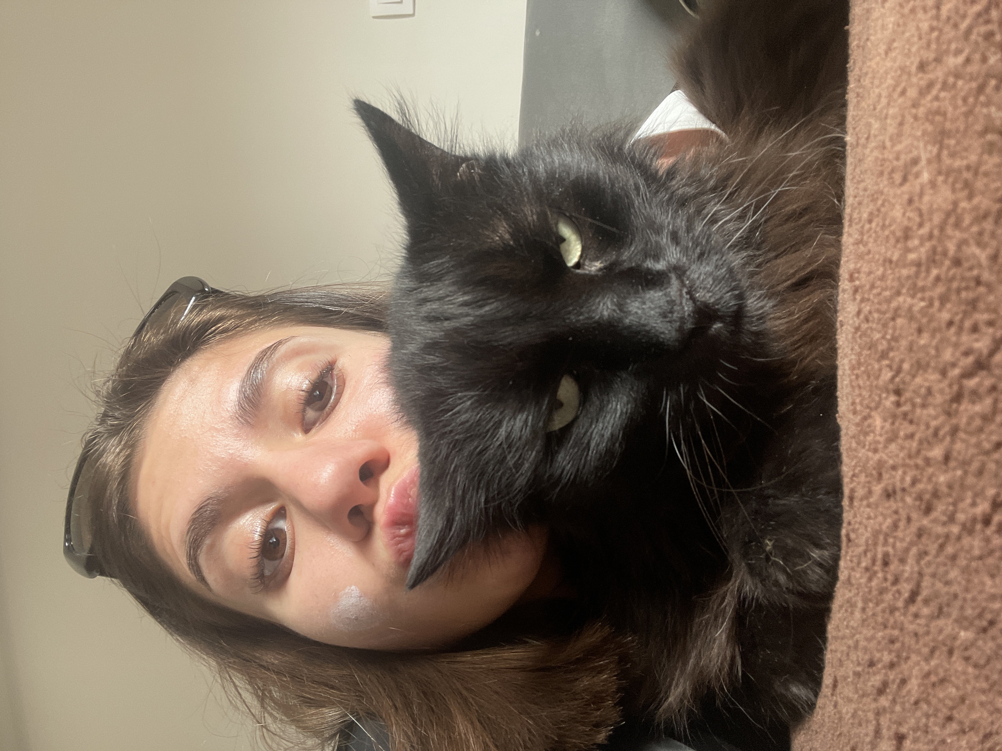The image displays a close-up, portrait-style photograph, currently oriented vertically with the subjects' heads positioned to the left. It features a young person and a large black cat posing intimately indoors in Sofia, Bulgaria.
The person, likely female, has medium-brown hair visible around her face and shoulder. She wears dark sunglasses pushed up onto her head. Her face is directed towards the camera, with her brown eyes clearly visible and her lips slightly pursed in a pouting expression. A small, light-colored smudge or product is present on her left cheek.
Snuggled closely against the person's face is a large, fluffy black cat. The cat's head dominates the right side of the frame, with its long black fur covering part of the person's nose and mouth. A prominent, pointed ear is visible at the top of its head, and long white whiskers extend from its muzzle. One eye, a striking greenish-yellow, is open and looks towards the camera.
The background includes a plain, light-colored wall behind the person's head, where a white electrical outlet or switch is visible. To the right of the cat, a textured brown surface, possibly a blanket or upholstered furniture, is present. The lighting suggests an indoor setting, and the overall mood is one of affection and playfulness between the person and their pet. No text is visible in the image.