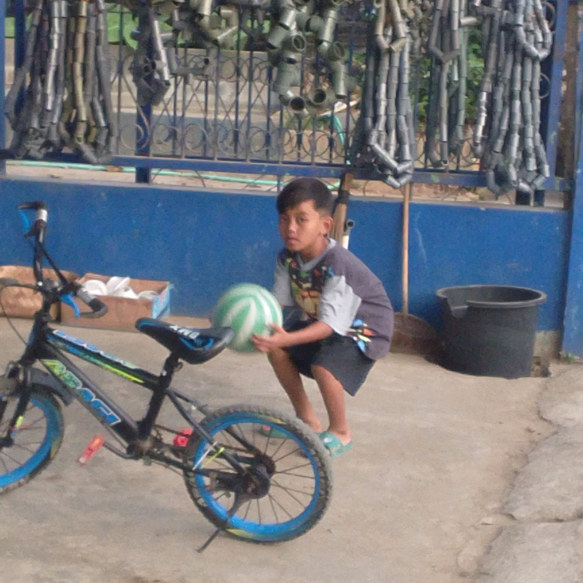 In Kroyo Lor, Indonesia, a young boy crouches on a concrete ground, holding a green and white striped ball. He is positioned to the left of a black bicycle with blue rims and neon green accents. The bicycle has a visible "ABACI" logo on its frame. Behind the boy and the bicycle is a bright blue wall, and further back, a metal fence adorned with numerous hanging pipes. To the right of the boy, a black plastic bucket sits on the ground. In the background, beyond the blue wall and fence, greenery is visible. The scene appears to be outdoors, possibly in a residential or communal area.