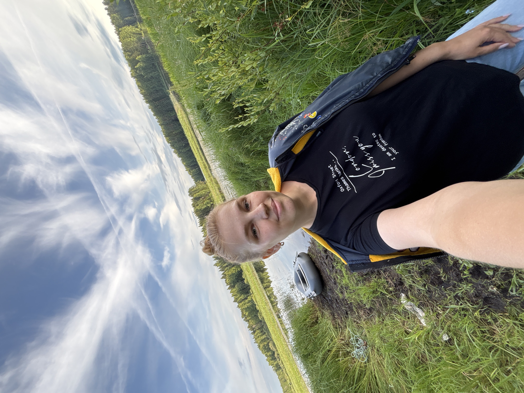 A young woman with blonde hair in a bun takes a selfie by a body of water, with trees and a blue sky with wispy clouds in the background. She is wearing a black t-shirt with white text that reads "Oxford Street Thames banks... London, I miss you. I am devoted to your freedom" and a dark jacket with yellow trim. In the water, a deflated inflatable boat is visible. The ground is muddy and covered in grass, with some litter. The location is Anteplevo (Okhotinskoe S/P), Russia.
