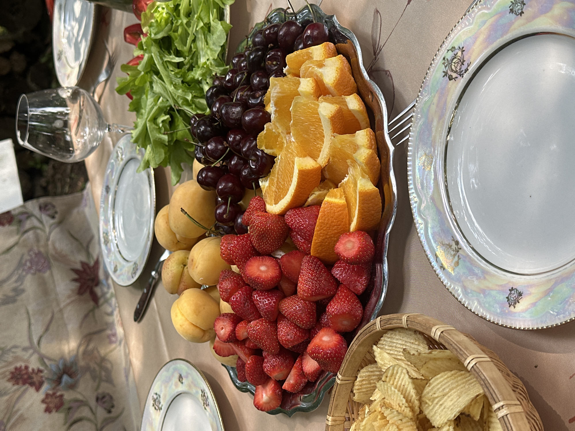 A close-up overhead shot shows a rustic outdoor picnic table laden with food. A silver platter is filled with strawberries, sliced oranges, apricots, and cherries. To the left, a basket overflows with potato chips. In the background, a table runner with a floral pattern is draped over the table, and a glass wine glass sits empty. The scene appears to be set in Козино, Russia, suggesting a leisurely meal in a natural setting.