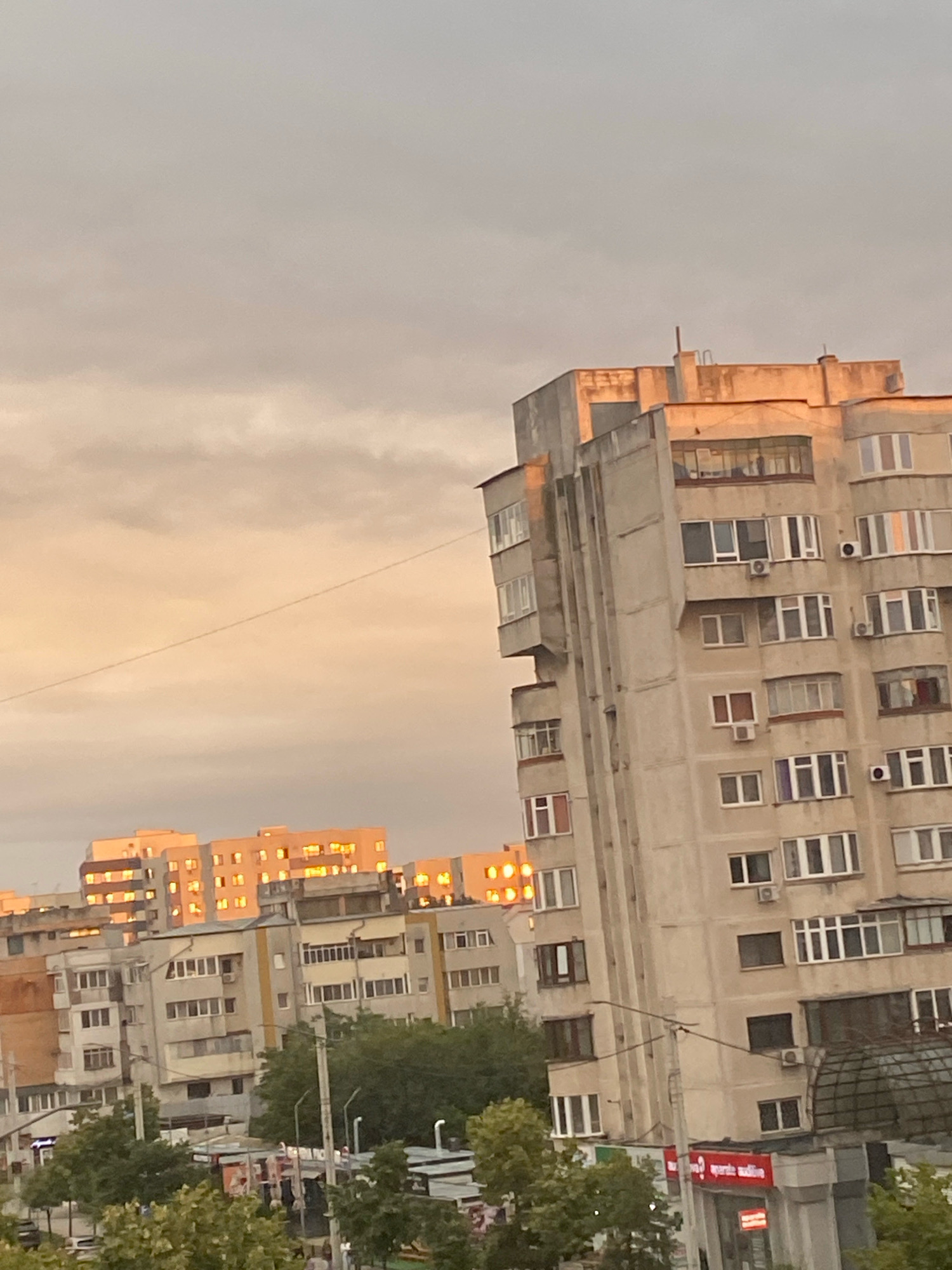The image depicts an urban cityscape in Iasi, Romania, during what appears to be either sunrise or sunset, given the warm, golden light illuminating the buildings and the sky. The foreground features a dense collection of apartment buildings, characterized by their concrete facades and numerous windows. Some of these buildings exhibit signs of wear and tear, with visible discoloration and balconies. In the mid-ground and background, more apartment blocks are visible, some catching the sunlight more directly, appearing brighter and more golden. The sky above is overcast with diffused clouds, tinged with orange and yellow hues. A few trees are visible at the bottom of the frame, suggesting the presence of green spaces within the urban environment. There are also some street-level elements, including what appear to be shops or businesses with signage, and streetlights. No individuals are clearly discernible. The overall impression is of a densely populated residential area bathed in the soft, atmospheric light of twilight.