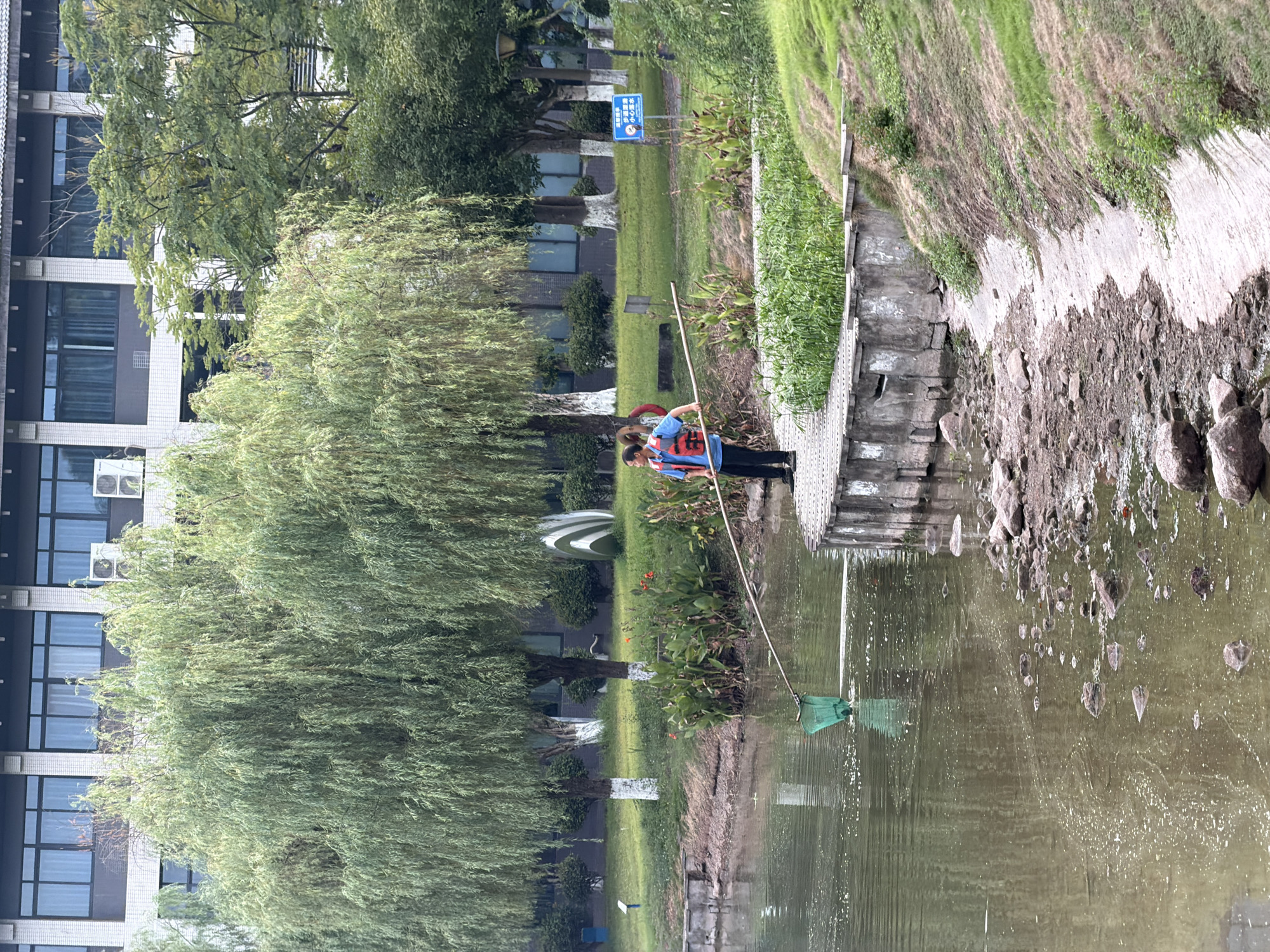In Jiangjin, China, a person wearing a blue shirt and a life vest is standing on a paved walkway beside a body of water. They are holding a long pole with a green net attached, which is submerged in the water. The person appears to be fishing or cleaning the water. The environment is natural, with trees, grass, and plants surrounding the water. A building with windows is visible in the background. The water's surface is rippled, and small white specks are scattered across it. The walkway is made of grey concrete, and rocks are visible along the edge of the water.
